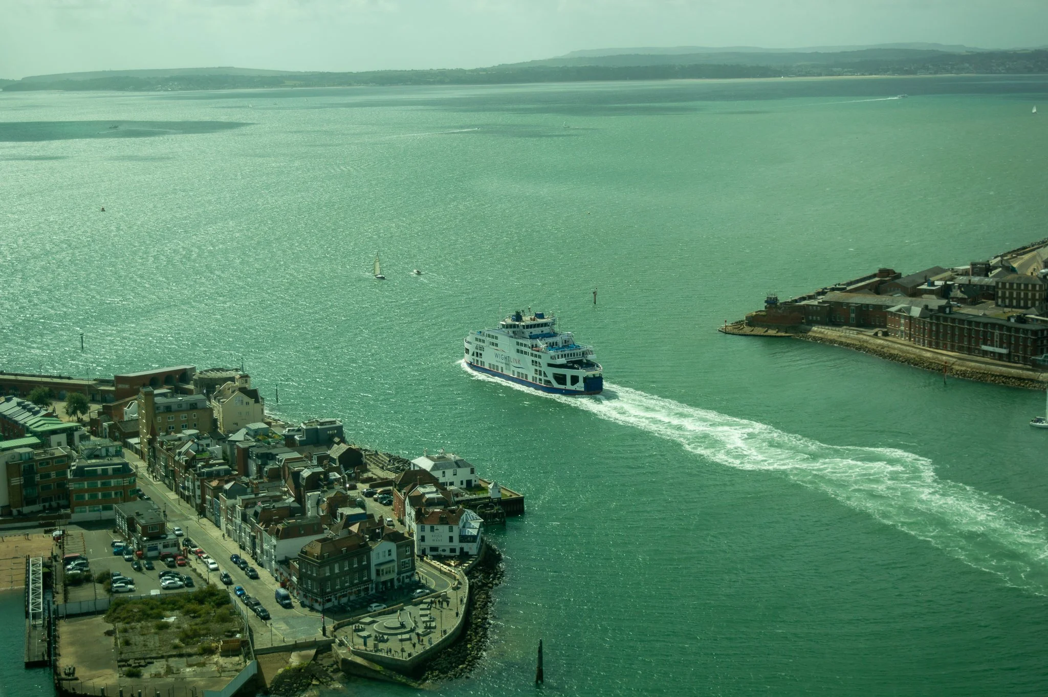 Aerial view of a harbor with a large white ferry boat labeled 'Wightlink' traveling through the water, alongside smaller boats and sailboats, with a cityscape of colorful buildings along the waterfront and rolling hills in the background.