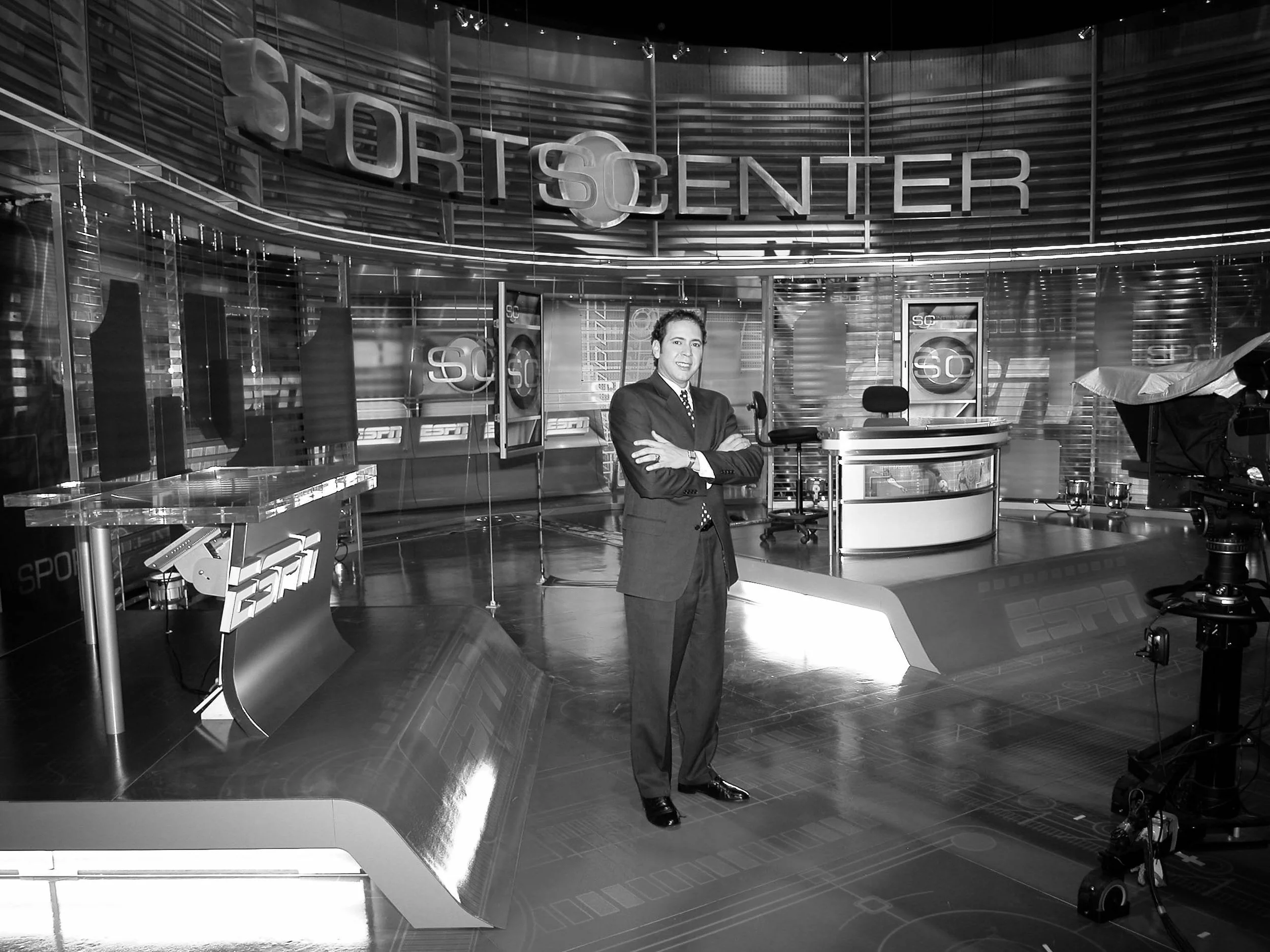 Studio news set with a man in a suit standing with arms crossed, behind a news desk, with projections and monitors displaying ESPN Sportscenter branding.