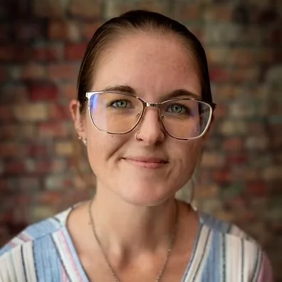 A woman with glasses and a striped shirt standing in front of a brick wall.