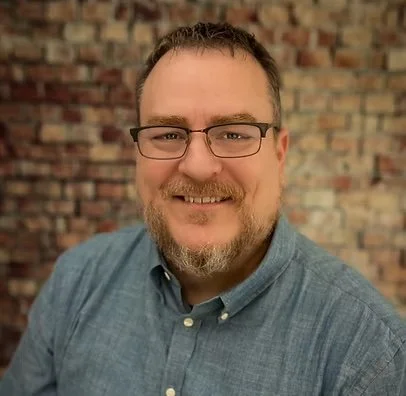 A man with glasses, a beard, and short hair smiling at the camera, wearing a blue button-up shirt with a brick wall in the background.