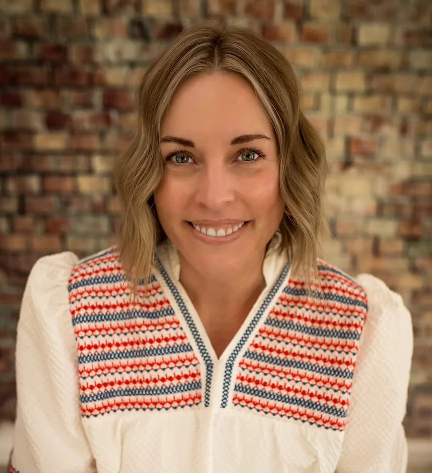 A woman with shoulder-length wavy brown hair and blue eyes, smiling, wearing a white sweater with a red, blue, and white patterned yoke, against a brick wall background.