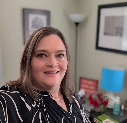 A woman with shoulder-length hair taking a selfie in a room decorated with wall art, a table with flowers, and a blue lamp.
