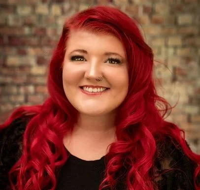 A woman with long, wavy red hair and a nose piercing, smiling against a brick wall background.
