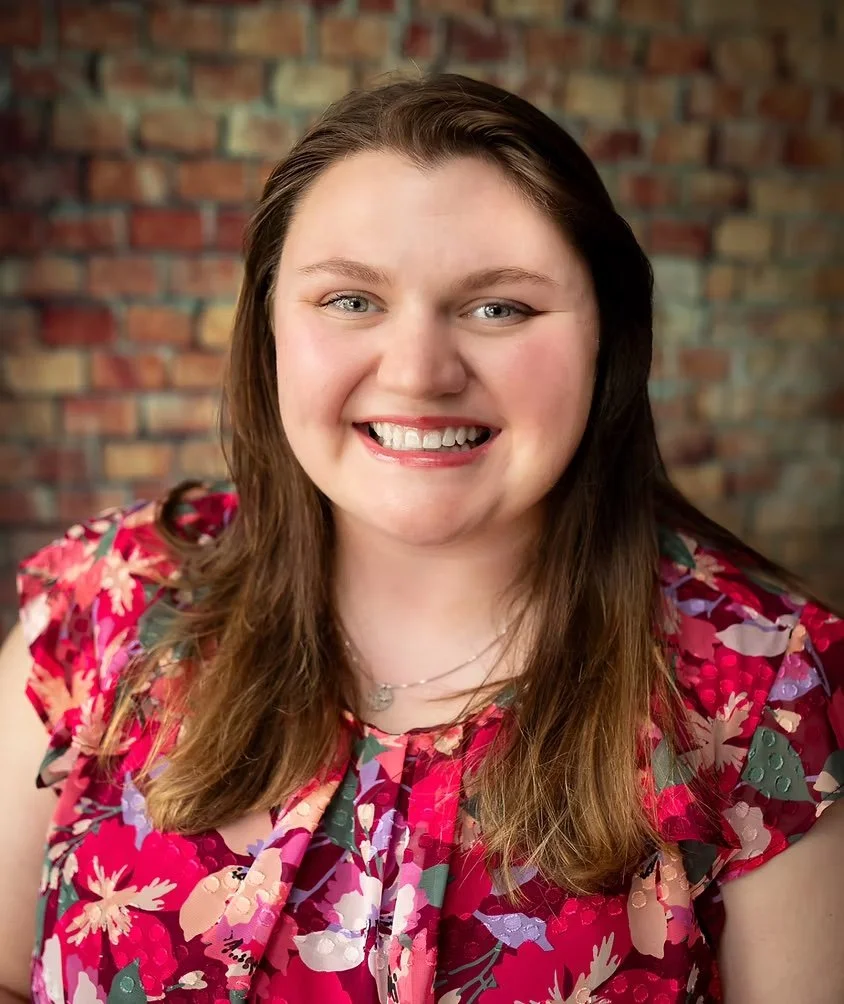 Close-up of a smiling woman with long brown hair, wearing a floral pink dress, in front of a brick wall.