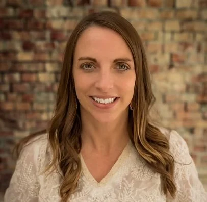 A woman with long, wavy brown hair smiling in front of a brick wall.