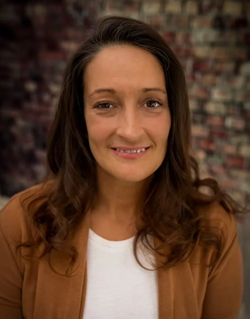 A woman with long, wavy brown hair smiling in front of a brick wall.