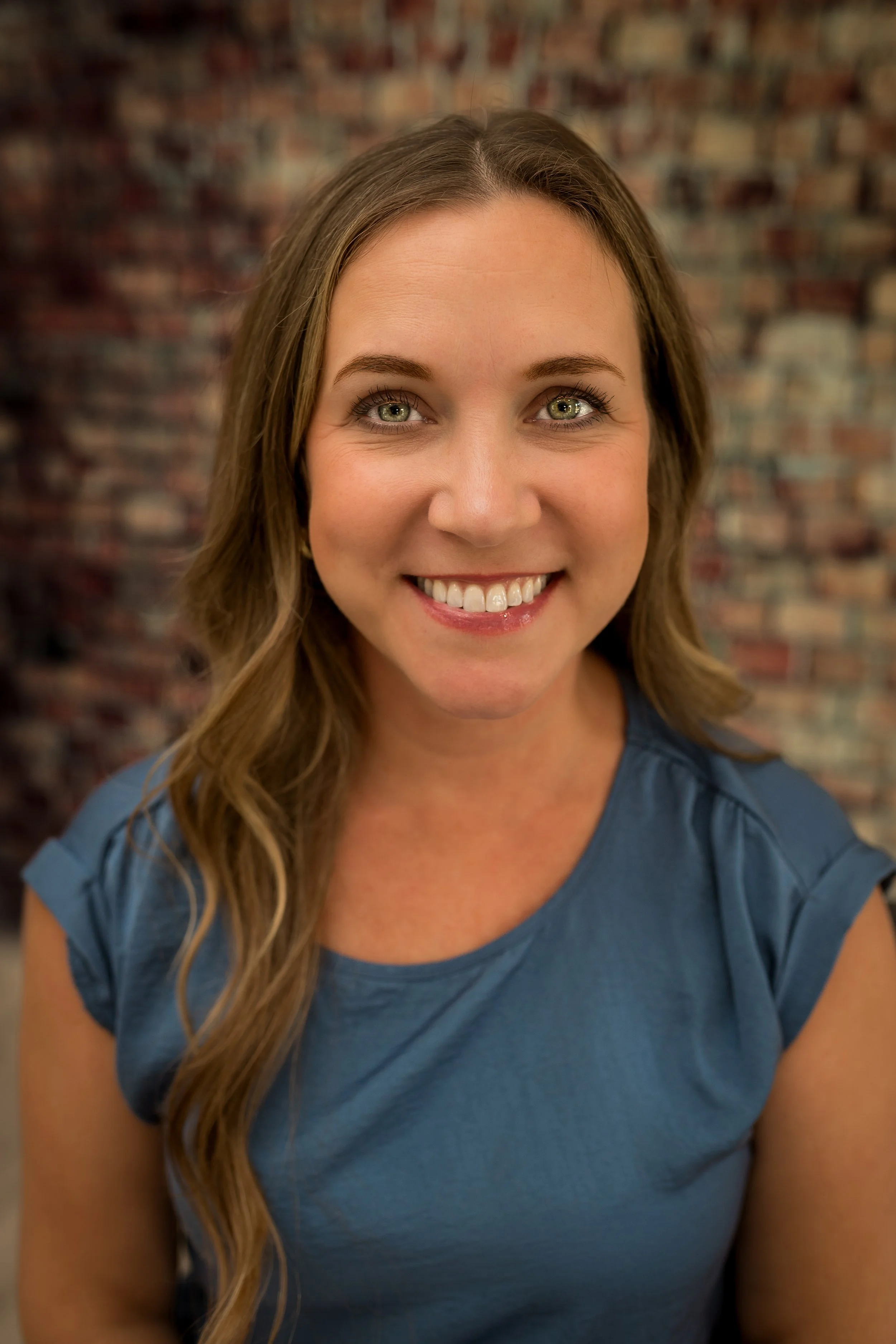 Close-up photograph of a smiling woman with long wavy hair, wearing a blue top, in front of a blurred brick wall background.