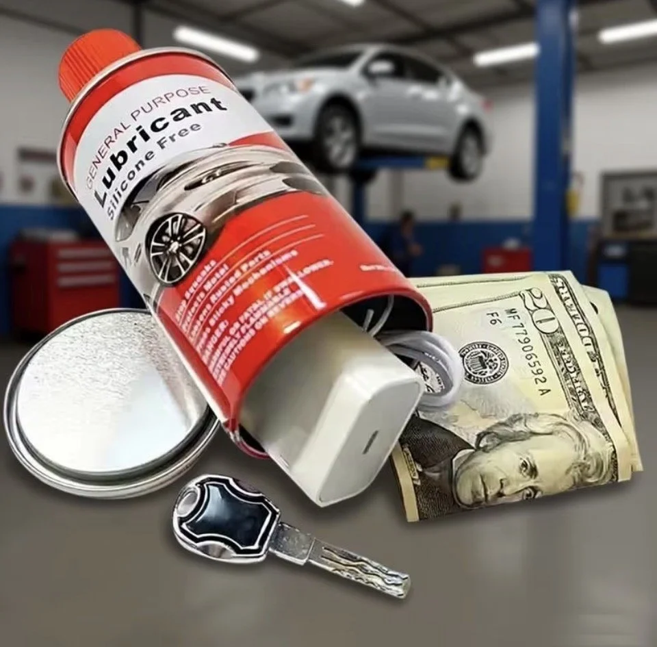 A can of silicone lubricant, a dollar bill, a key, and a small round container are on a car workshop table with a vehicle lifted in the background.