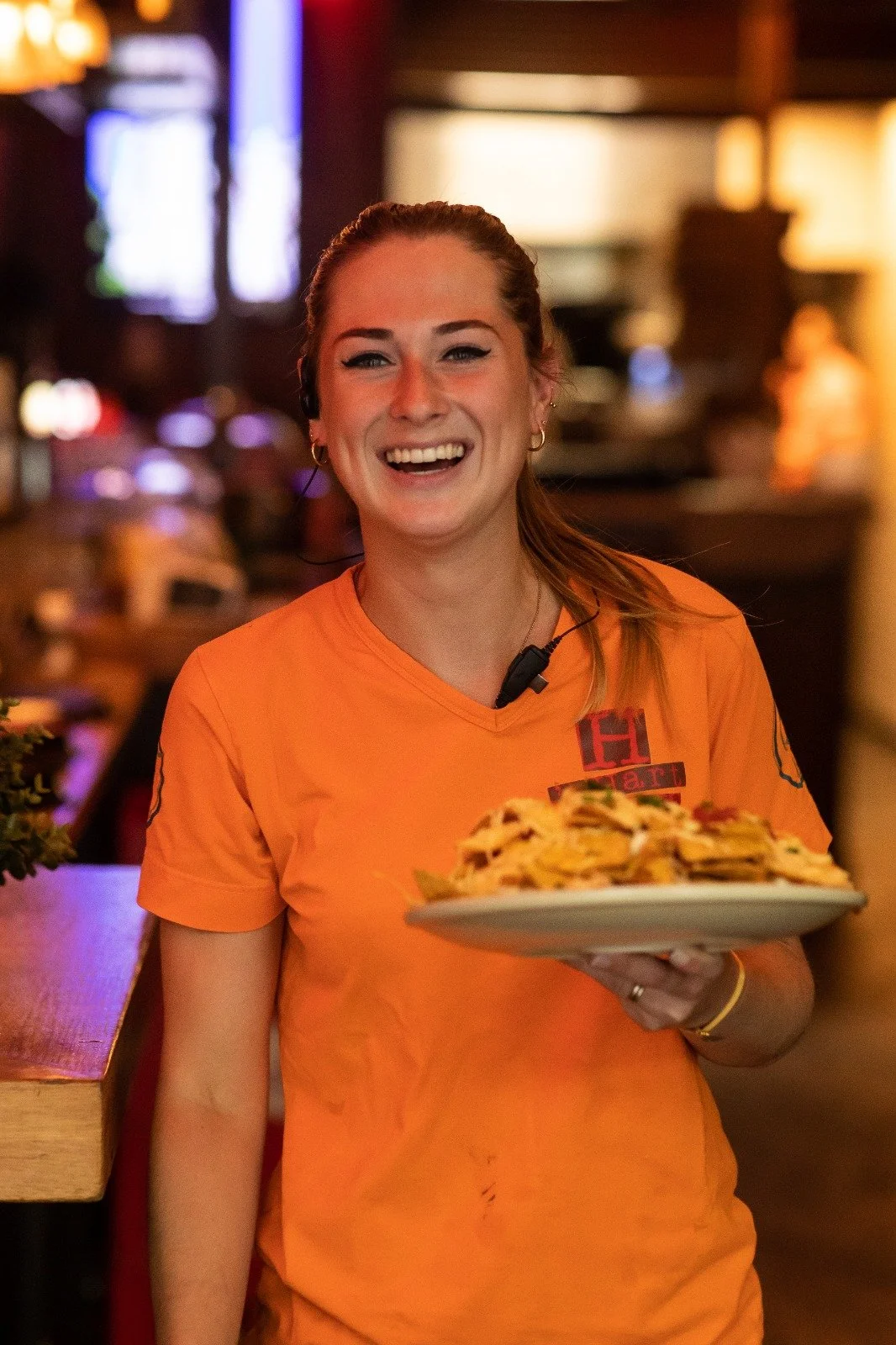 A smiling woman wearing an orange shirt holding a plate of food in a restaurant setting.