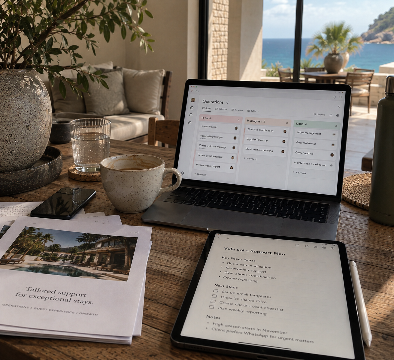 A workspace on a wooden table with a laptop displaying a project management app, a tablet with a support plan, a cup of coffee with coffee stains, a glass of water, a smartphone, papers with a printed image of a resort, and a large potted plant. In the background, there is a view of the ocean and a balcony with outdoor furniture.