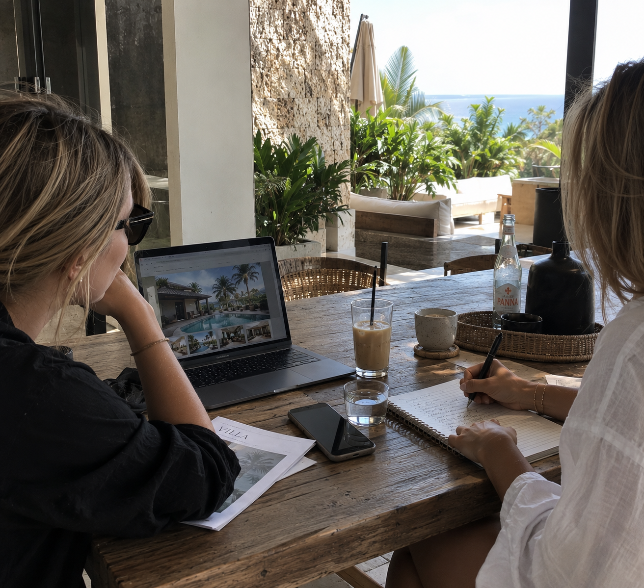 Two women sit at a rustic wooden table in an open-air café with tropical plants, looking at a laptop displaying a poolside resort. One woman is taking notes in a notepad, while the other is resting her head on her hand. There are drinks, a smartphone, a water bottle, and decorative items on the table. The background shows patio furniture and lush greenery near the ocean.