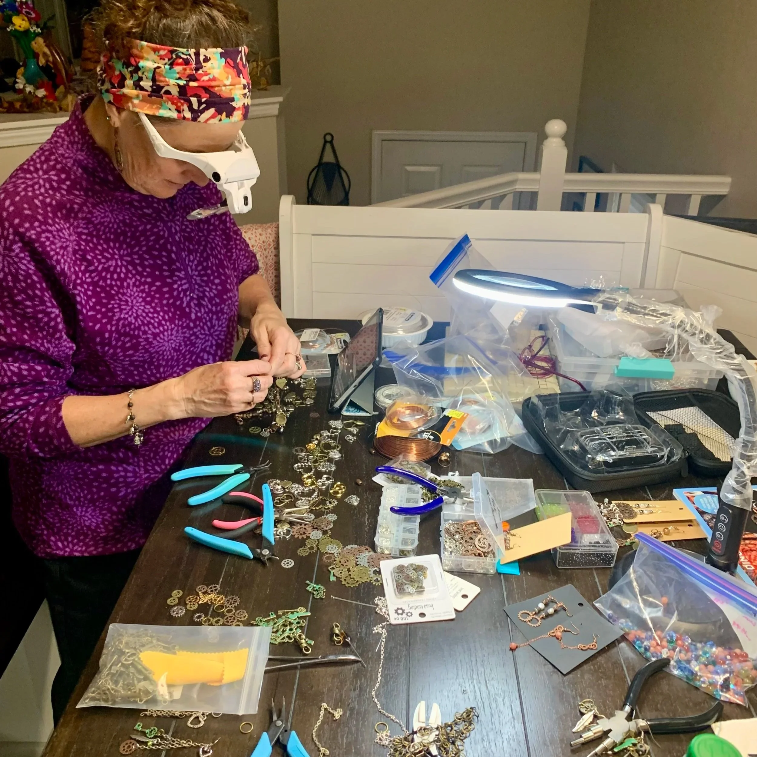 Terrie wearing a colorful headband and purple shirt is working at a table covered with jewelry-making supplies, including chains, charms, pliers, and containers of beads and findings.