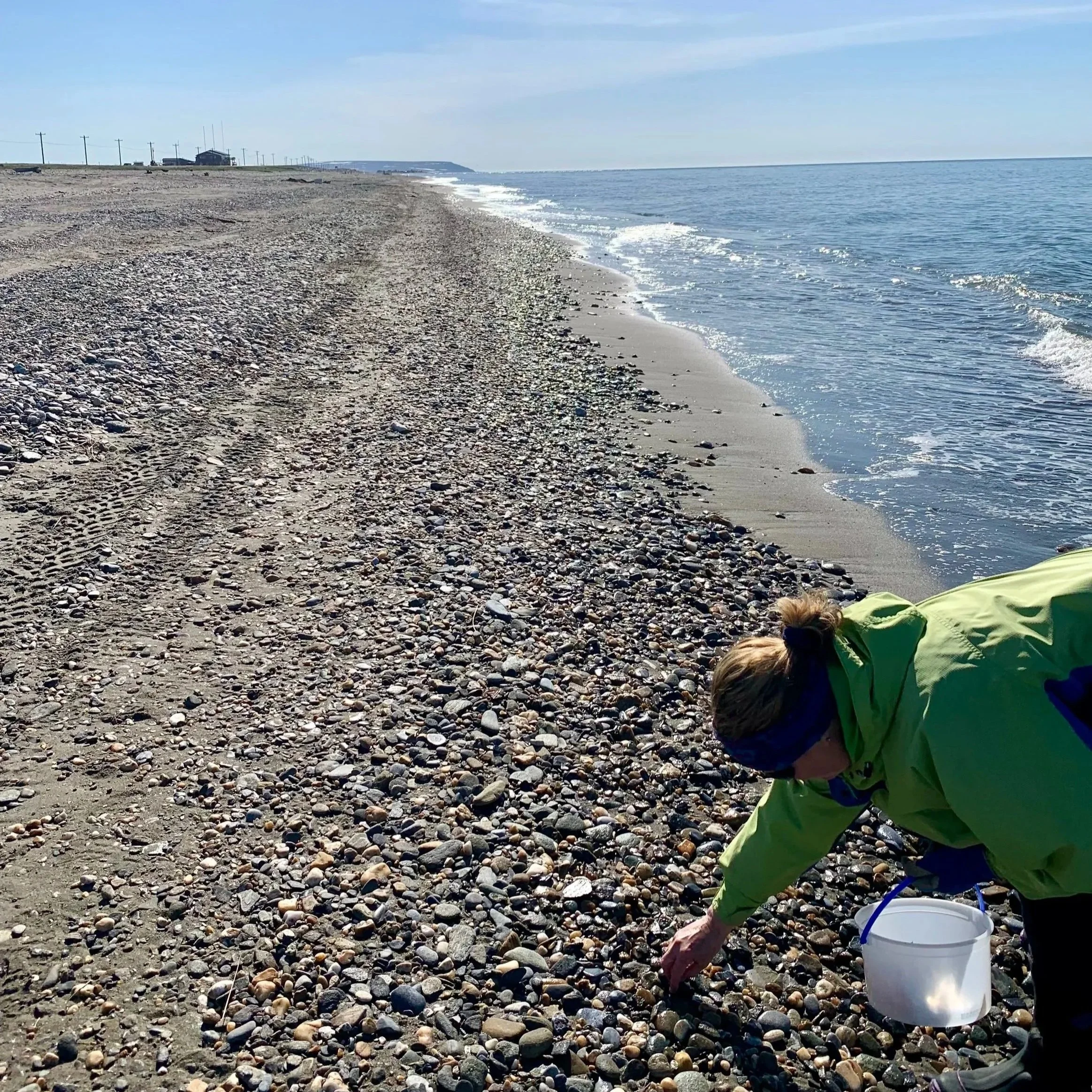 Terrie in a bright green jacket bending on a rocky beach, collecting sea glass into a white bucket with a blue handle, with the ocean and a partly cloudy sky in the background.