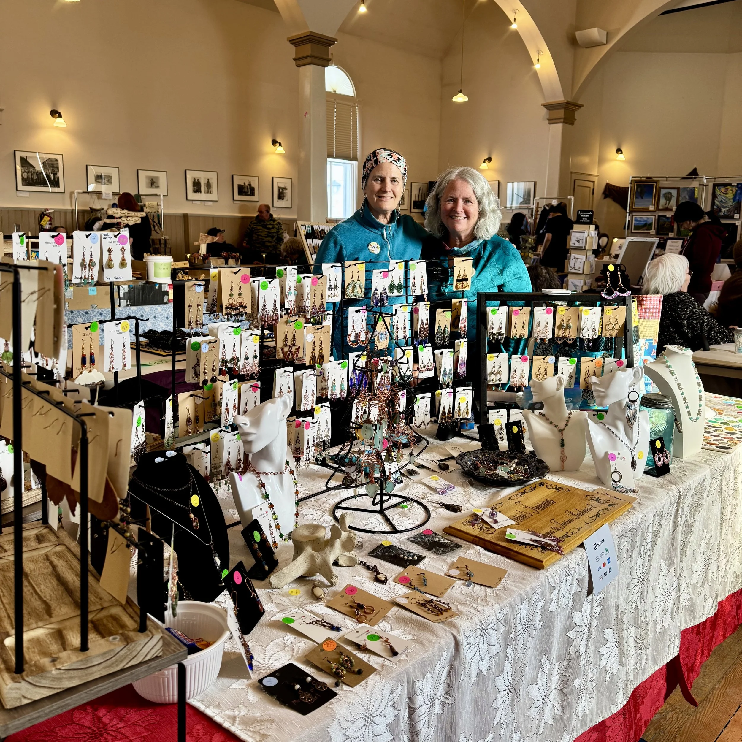 Terrie and her sister behind their display table at an indoor craft fair, showcasing earrings, necklaces, and other handmade pieces.