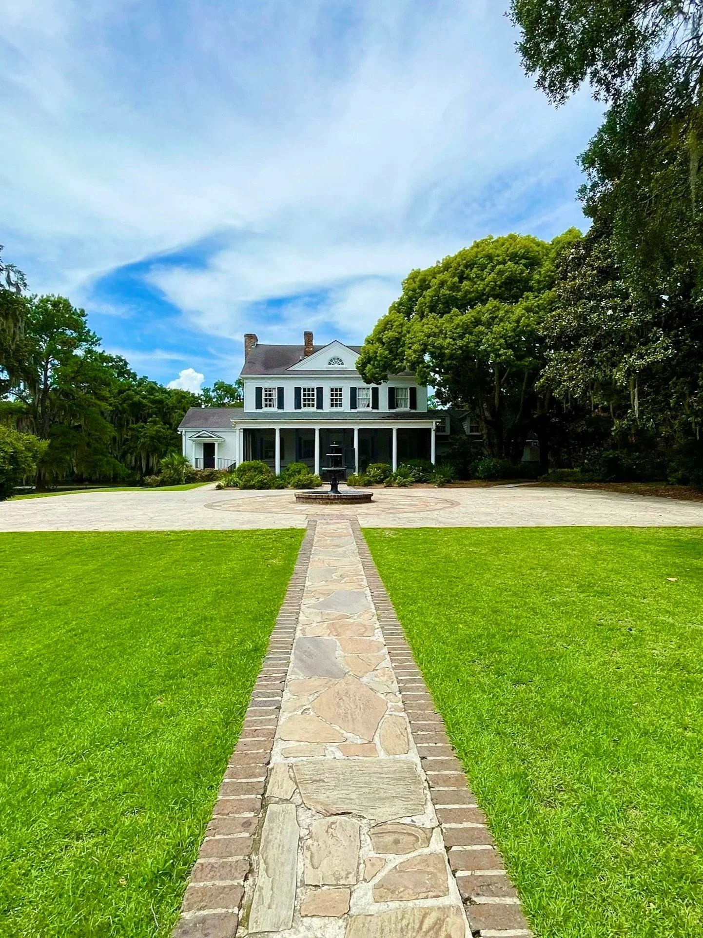 A stone walkway leads to a large white house surrounded by green trees and a well-manicured lawn.