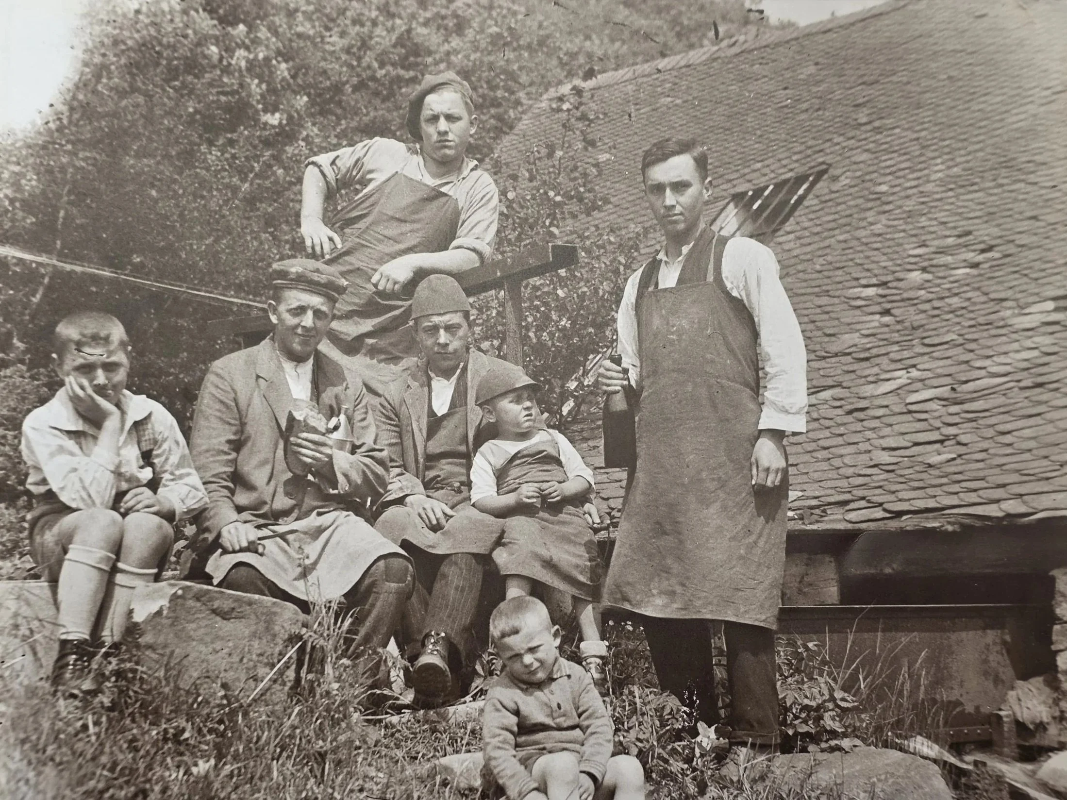 Schwarz-Weiß-Fotografie einer Gruppe von Menschen im Freien, teilweise sitzend, teilweise stehend, vor einer Hauswand mit Dach. Einige tragen traditionelle oder alte Kleidung, einige haben Hüte. Es scheint sich um eine historische oder alte Aufnahme zu handeln.