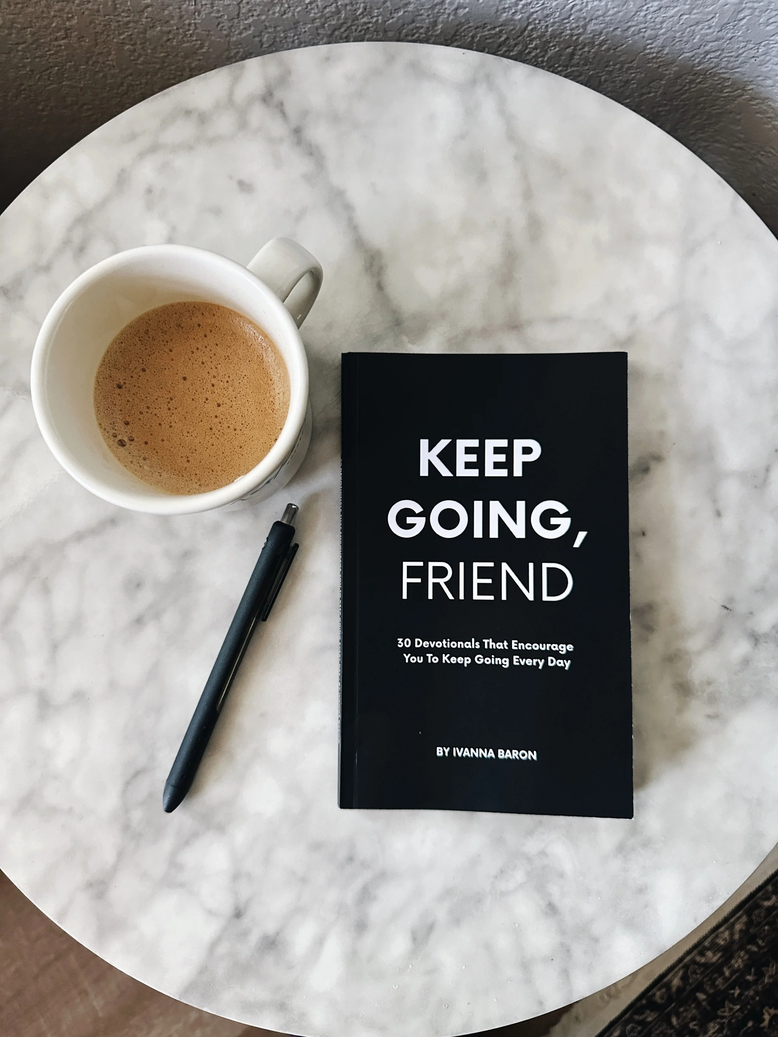 A white mug of coffee, a black pen, and a book titled 'Keep Going, Friend' on a white marble table.
