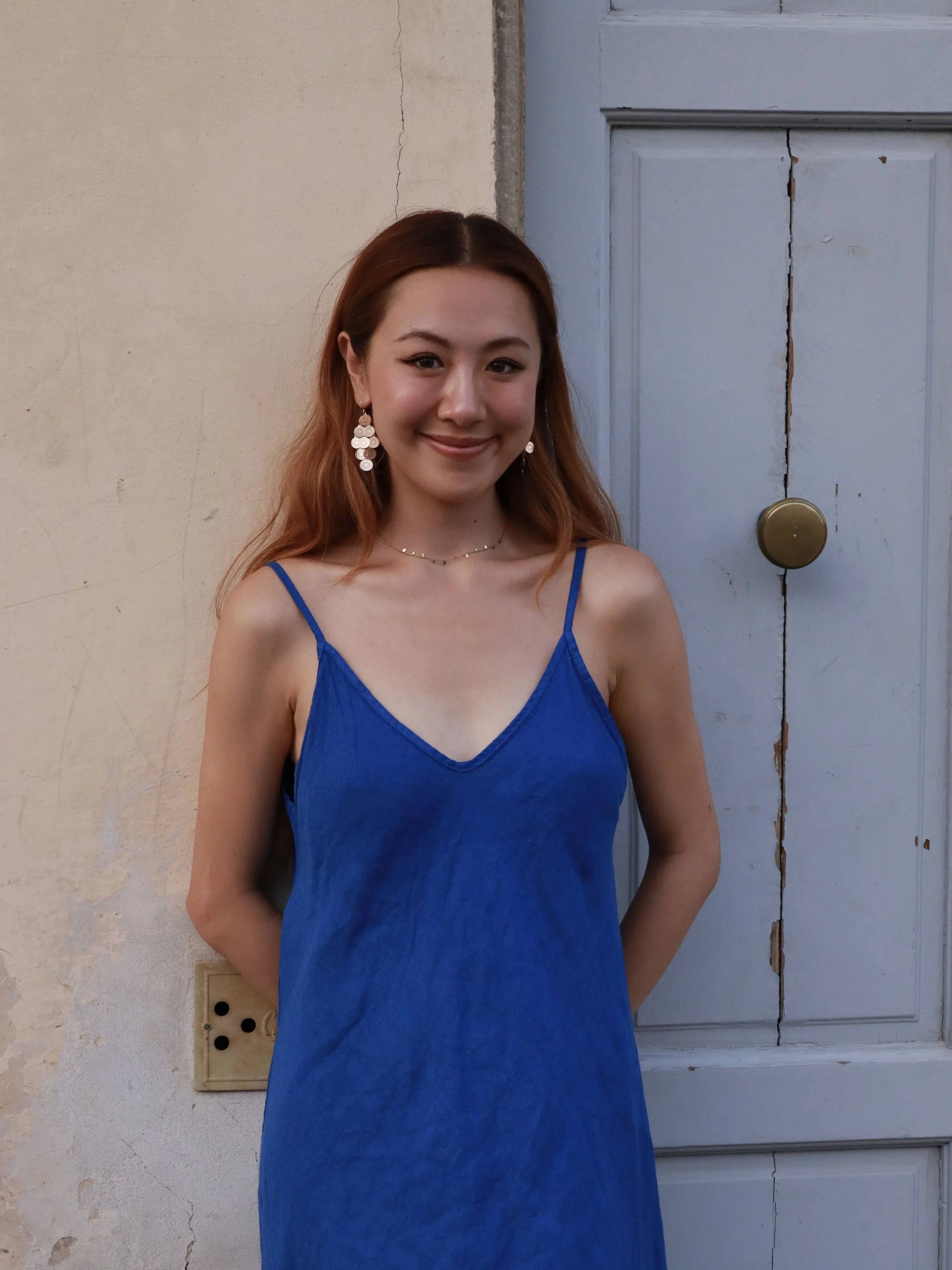 A young woman with reddish-brown hair, wearing a blue spaghetti strap dress and large dangling earrings, standing against a beige wall and a light blue wooden door with visible cracks and a brass doorknob.