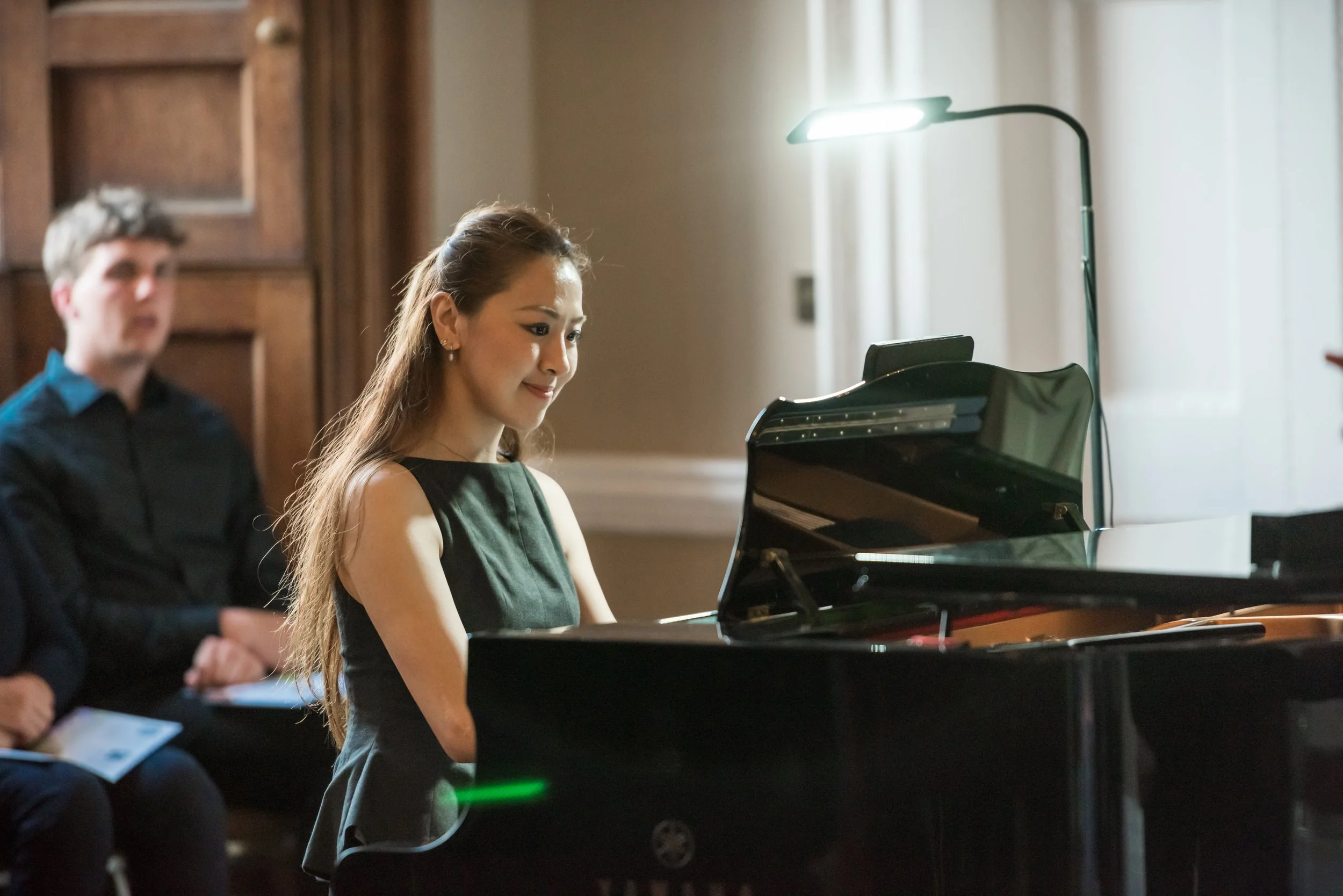 A woman playing a grand piano during a music performance, with a man sitting behind her, holding a sheet of music in a well-lit, wooden-paneled room.