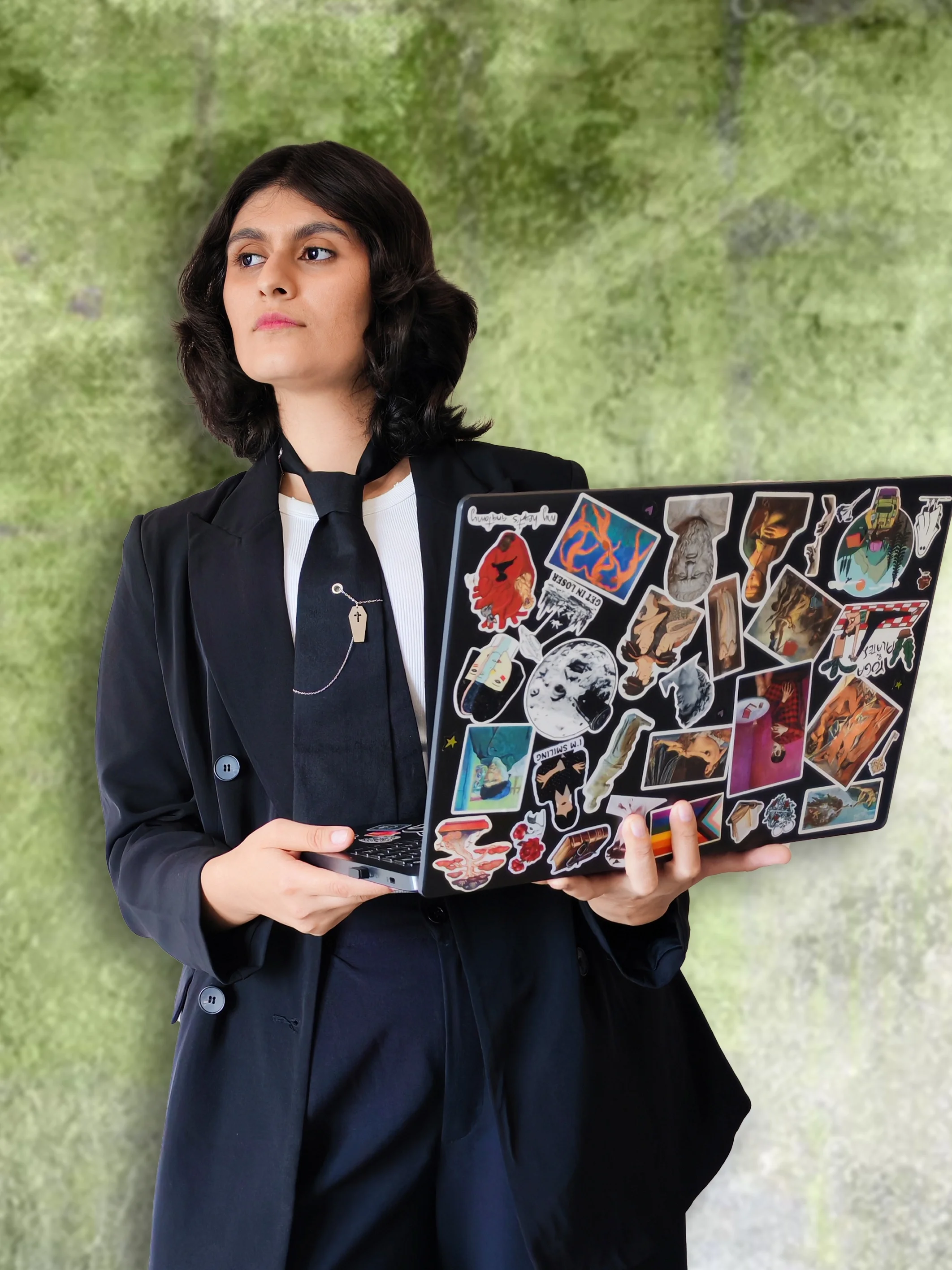 A woman in a black blazer and tie holding a laptop covered with various colorful stickers, standing outdoors with a blurred green background.