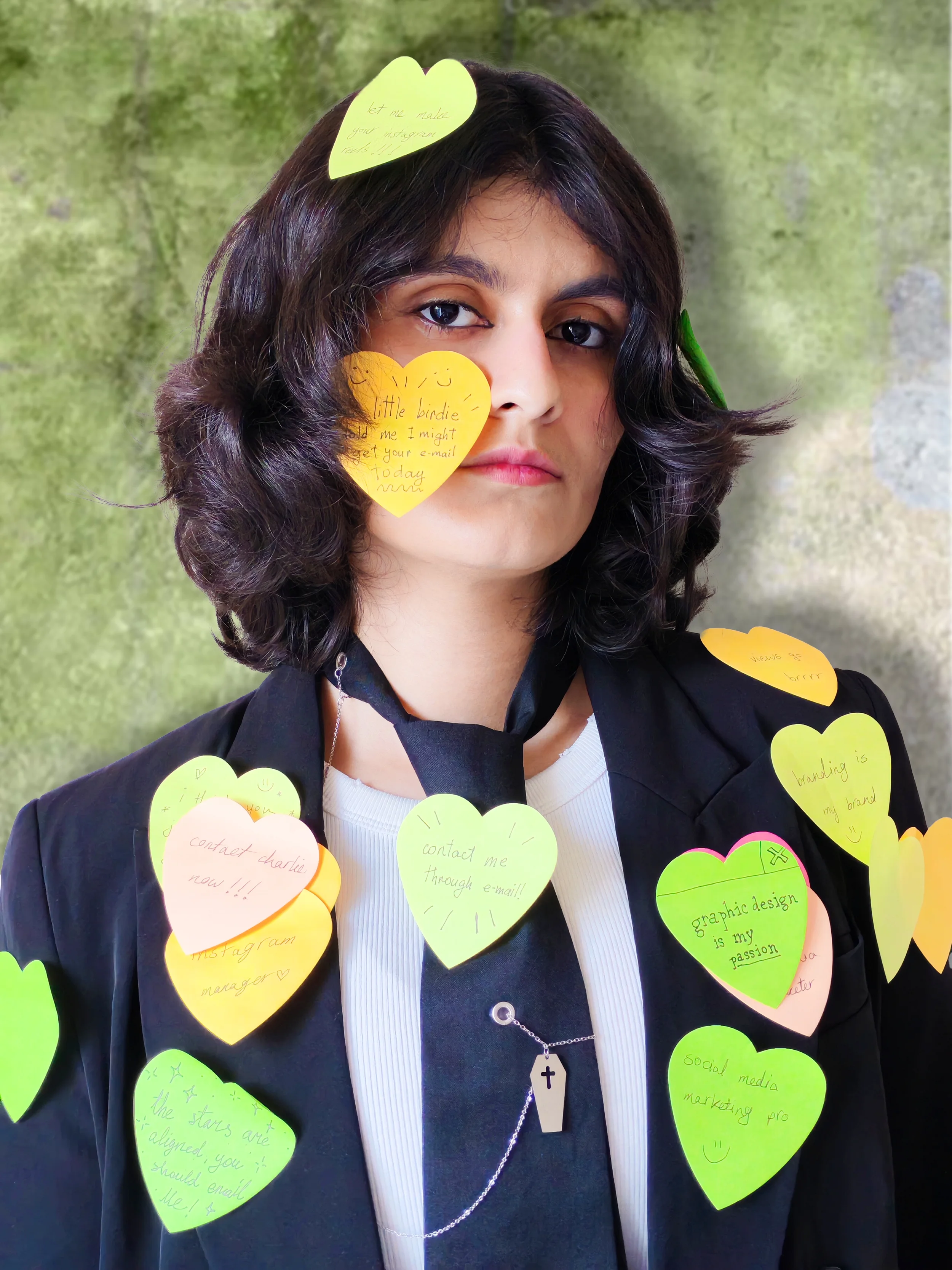 A young woman with dark, wavy hair wearing a black blazer and a white top, adorned with numerous yellow and green heart-shaped notes with handwritten messages on them. The notes include phrases about contacting her via email, social media marketing, graphic design passion, and other friendly notes. The background is blurred green.