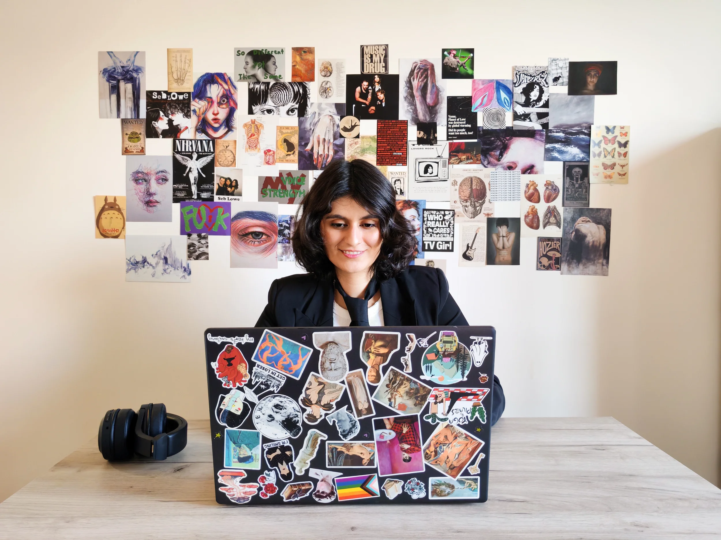 A person with shoulder-length dark hair and a black jacket sitting at a desk with a laptop that is covered in various stickers, and a black headphone is placed on the desk. Behind them, there is a collage of artwork, photos, and posters on the wall.