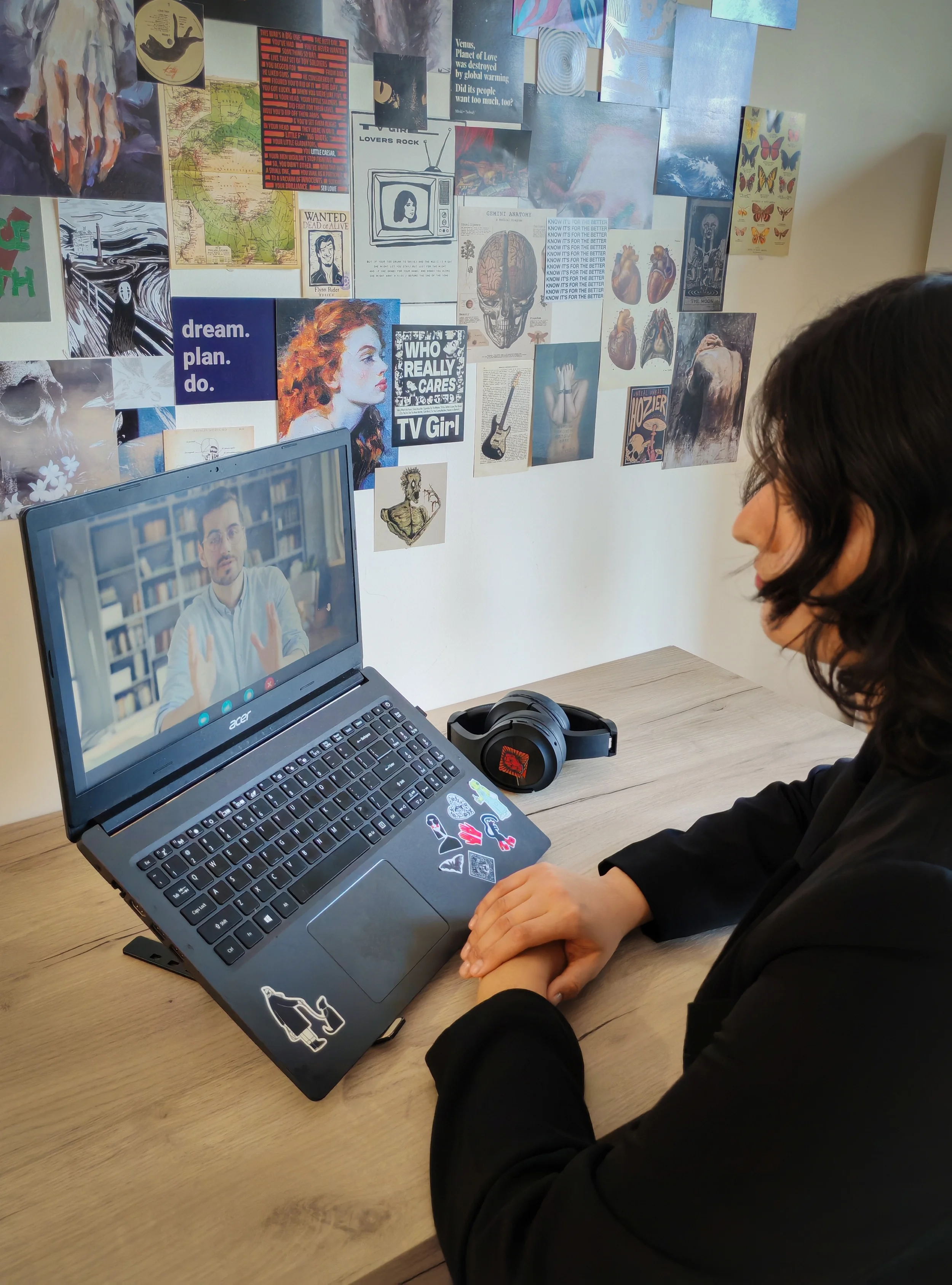 A person sitting at a desk using a laptop in front of a wall decorated with various posters, illustrations, and artwork, with a pair of headphones placed on the desk.