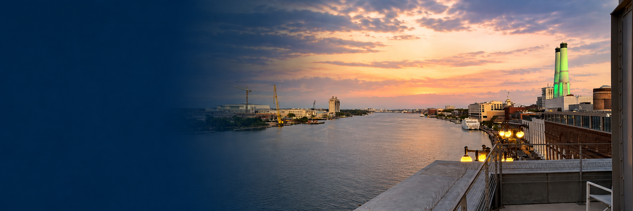 A city skyline at sunset beside a river, with buildings, boats, construction cranes, and a factory with green-lit smokestacks, viewed from a terrace with outdoor lights and a railing.