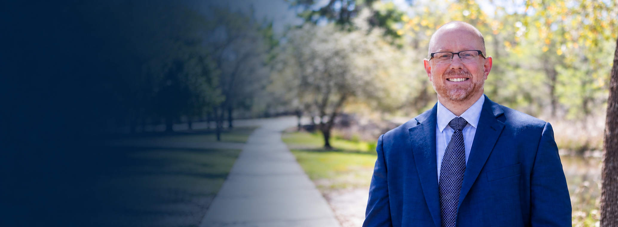 A smiling man in a blue suit, glasses, and a tie standing outdoors on a sidewalk with trees and greenery in the background.