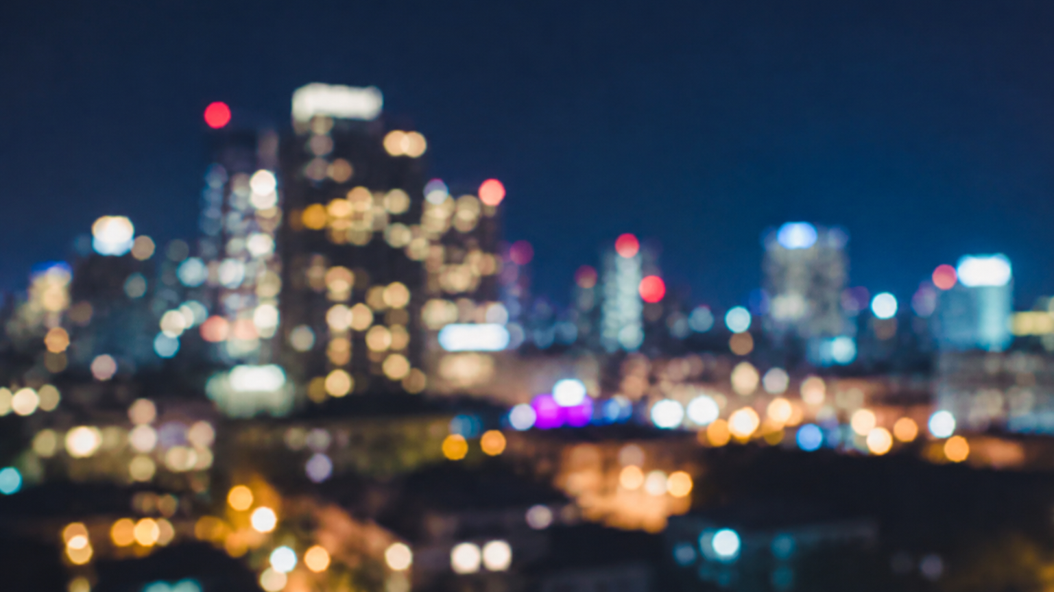 Blurred city skyline at night with colorful lights and tall buildings.