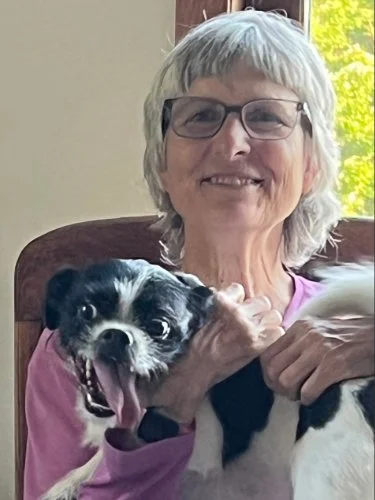 An elderly woman with short gray hair, glasses, and a pink shirt smiling while holding a black and white dog.