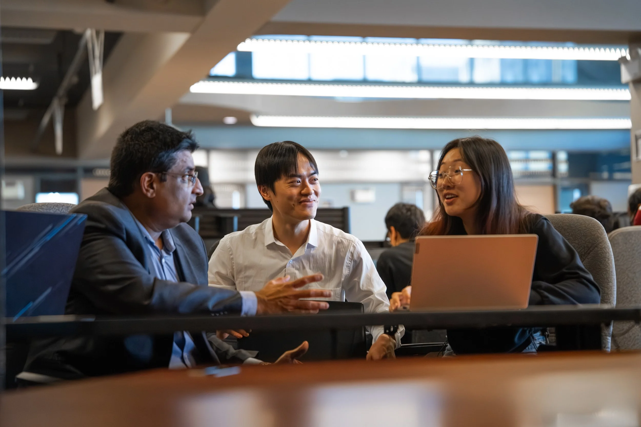 Three people engaged in a conversation at a table in a modern office or coworking space, with others working in the background.