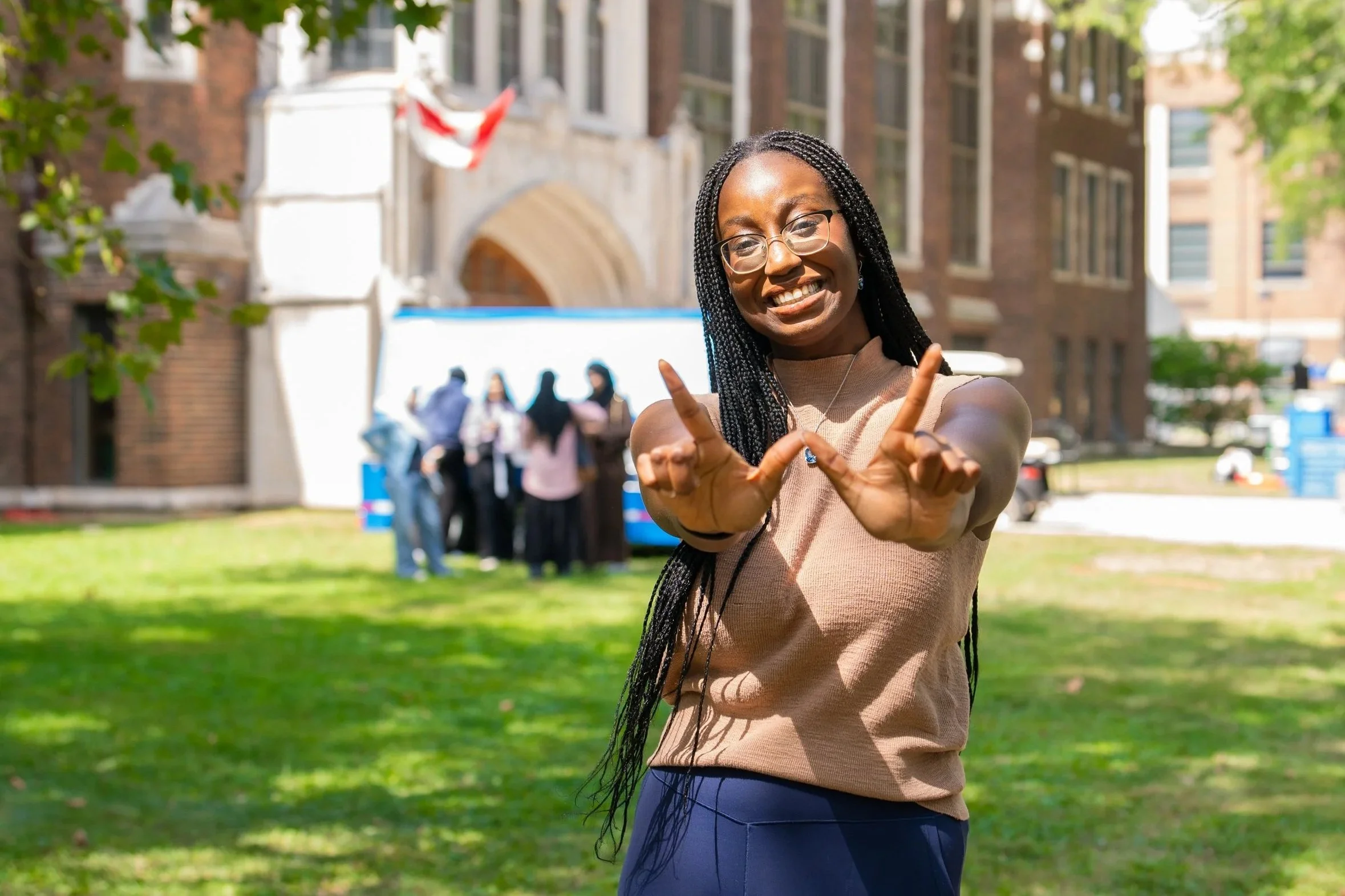 Smiling woman with glasses and long braids making a hand heart gesture outdoors in front of a group of people and a building.