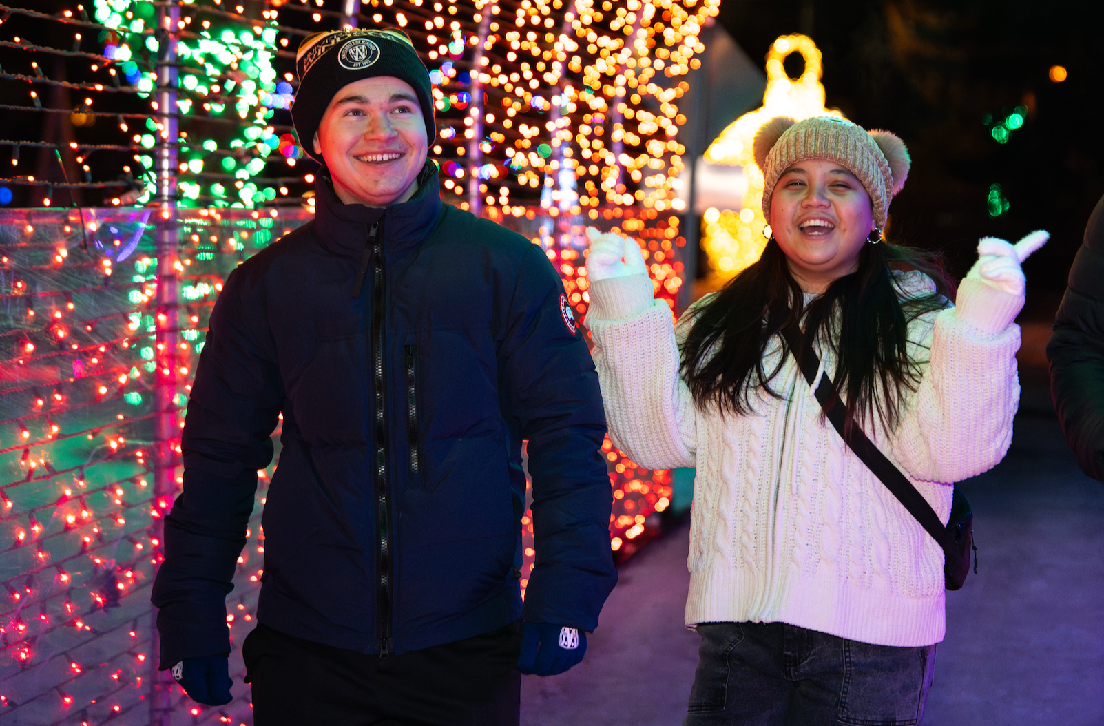 A smiling man and woman enjoying Christmas lights display at night. The man is wearing a black jacket and beanie, while the woman has on a beige beanie with pom-poms and a white knit sweater. Lighting decorations with multicolored lights are in the b