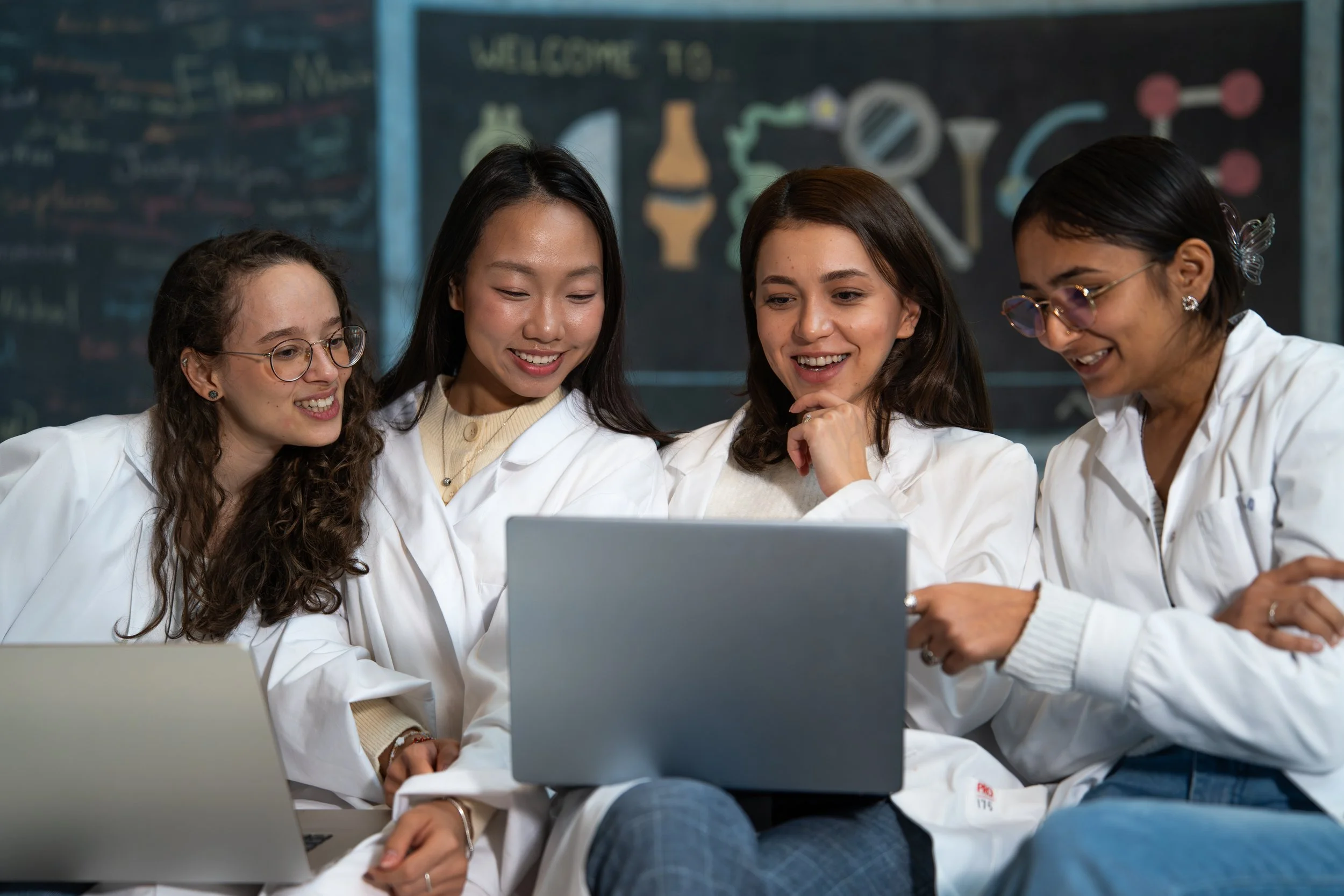Four women in white lab coats gathered around a laptop, smiling and looking at the screen in a science or educational setting.