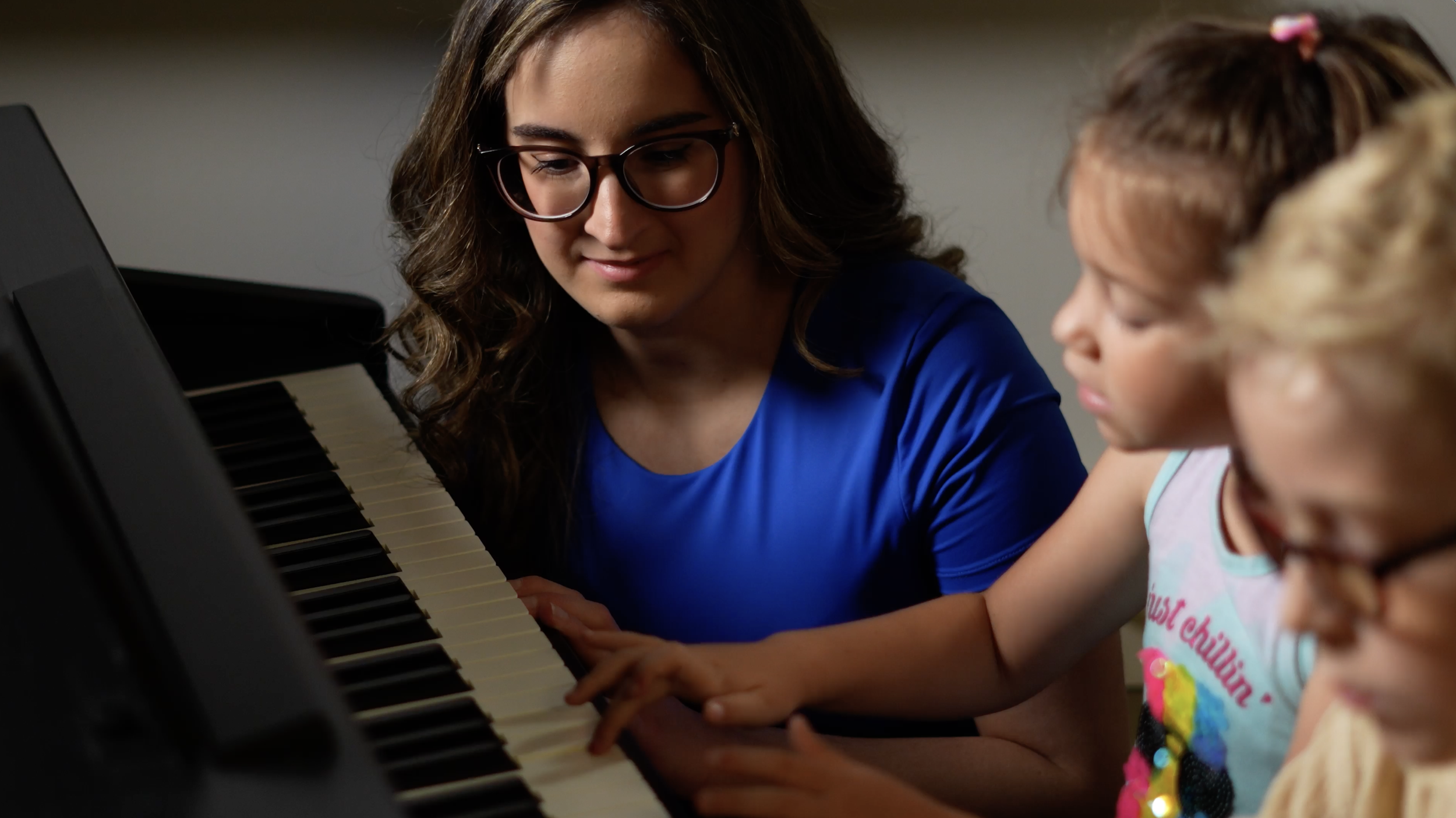 A woman with glasses playing the piano with two young girls, one of whom is looking at the keys, in an indoor setting.