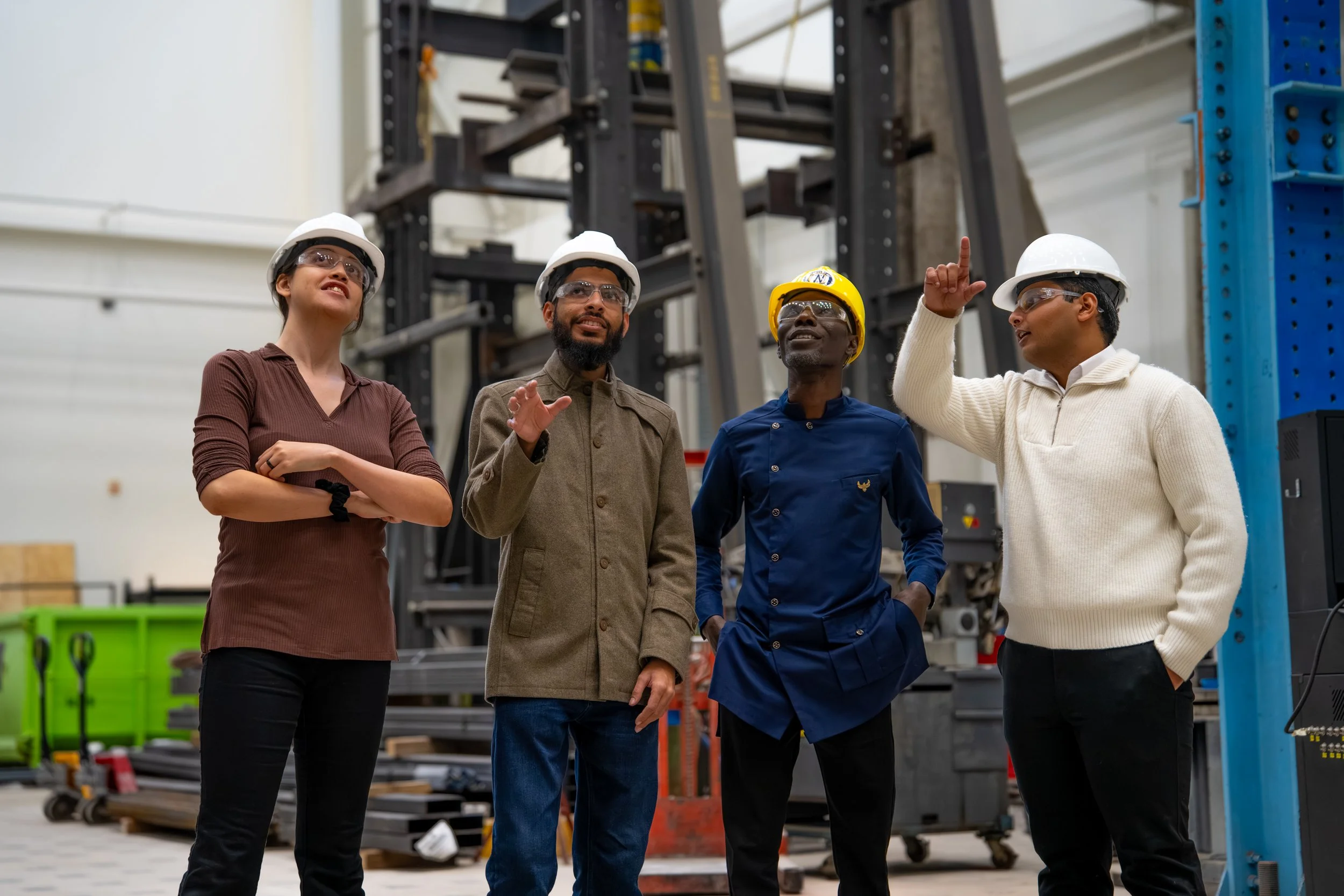 Four workers in safety helmets and glasses standing in an industrial warehouse near equipment, with one person explaining and others listening.