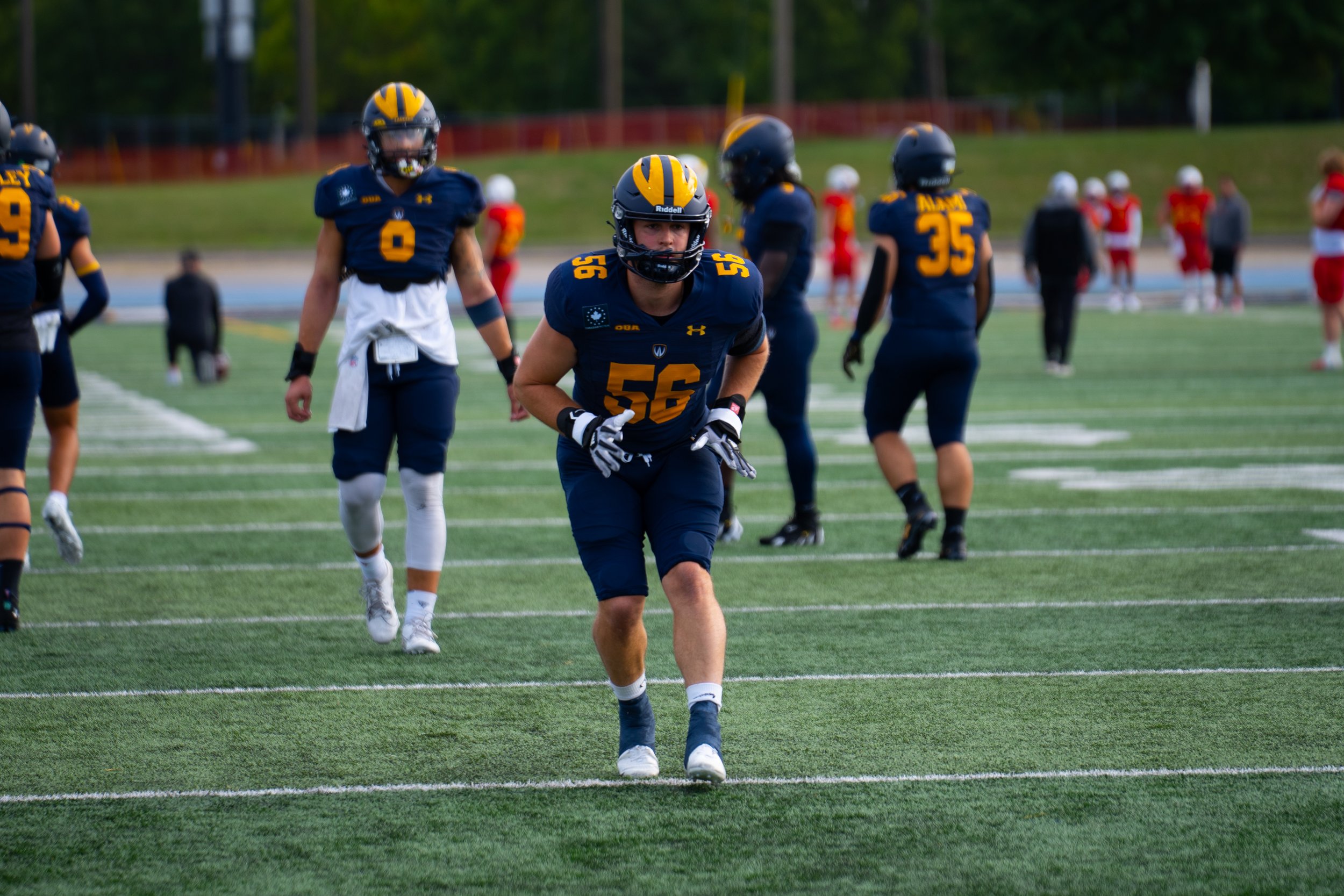 Youth football players in dark blue uniforms with yellow numbers and helmets on a field during a game. One player is in the foreground, crouching and focused, while others stand behind him. Players are on a green turf field with white yard lines, wit