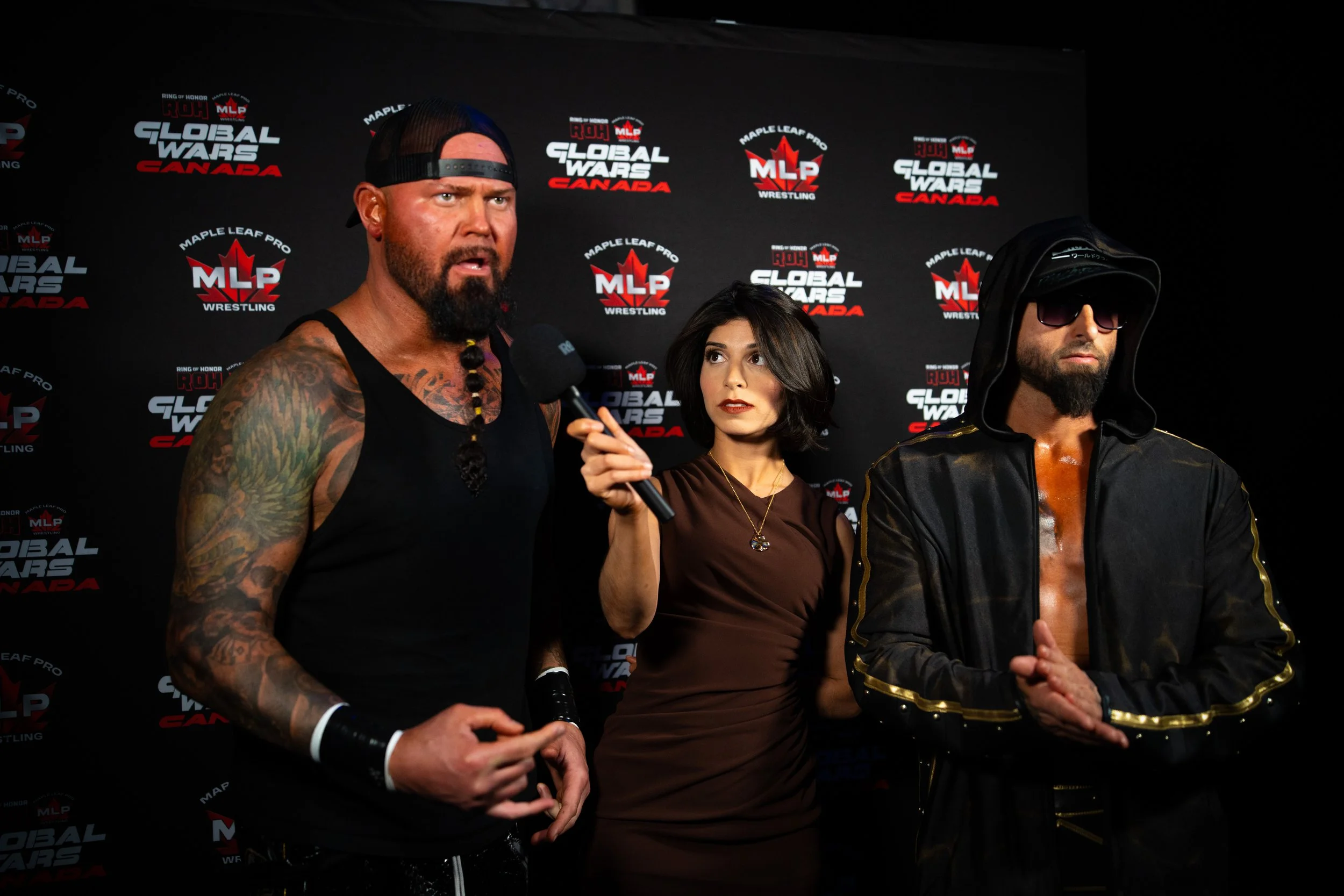 Three people at a press event with a black backdrop displaying logos for 'Global Wars Canada' and 'MLP Wrestling'. The man on the left has tattoos, a beard, and is wearing a black tank top and a backwards black cap. The woman in the center has shoulder-length dark hair and is holding a microphone, wearing a brown top. The man on the right wears sunglasses, a hat with a wide brim, and a black jacket with gold trim.