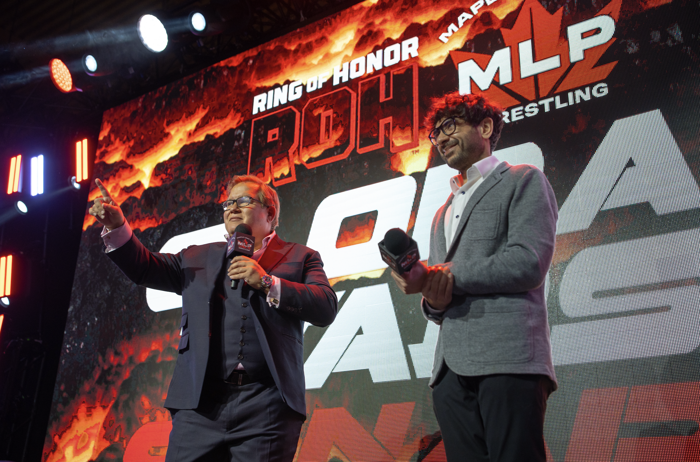 Two men standing on stage in suits holding microphones, with a large digital screen behind them displaying a logo and the words "Ring of Honor" and "MLW Wrestling," with fiery background imagery.