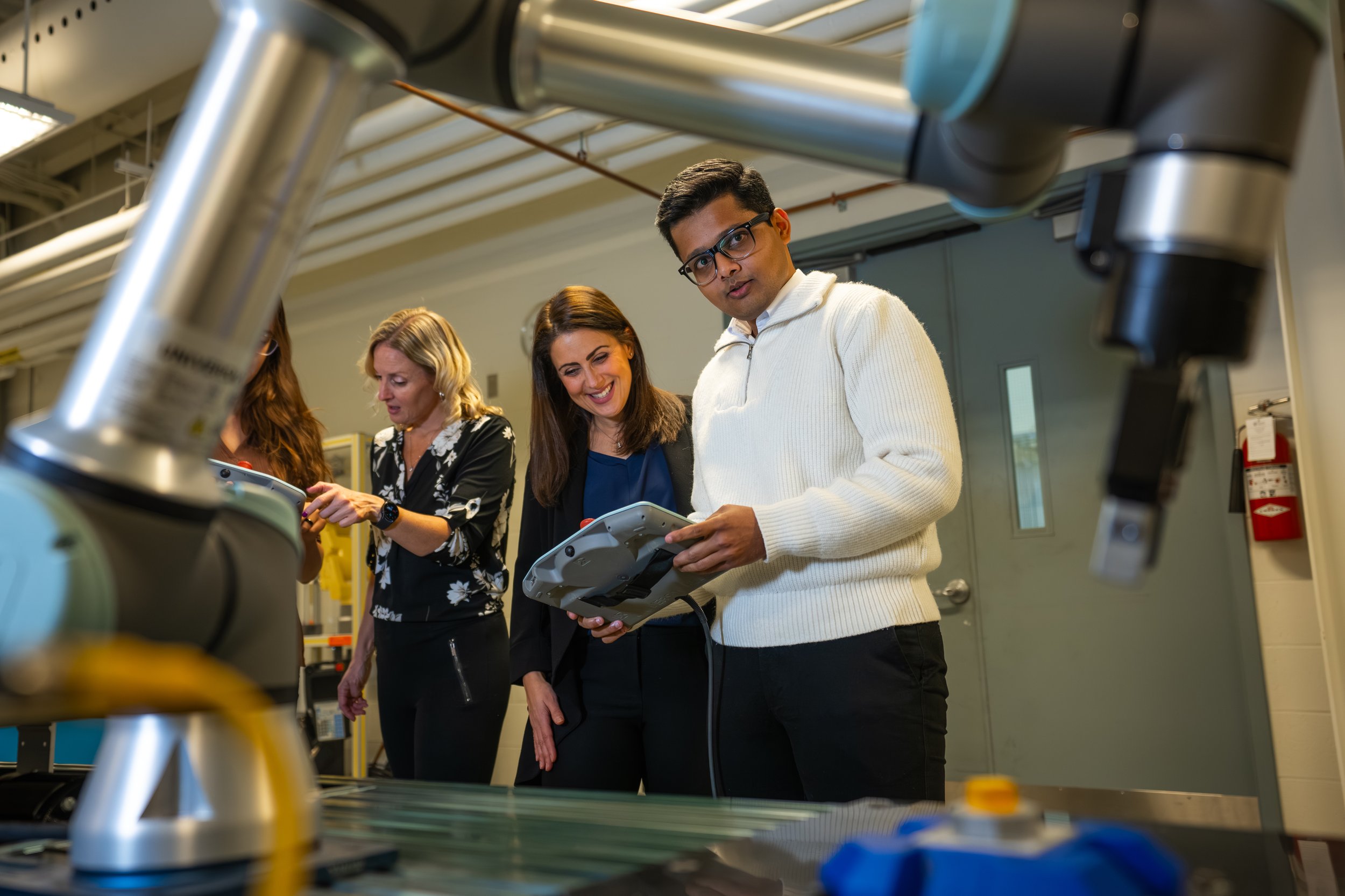 Group of five people in a science or engineering lab examining equipment, with metallic machinery in the foreground and a yellow safety box in the background.