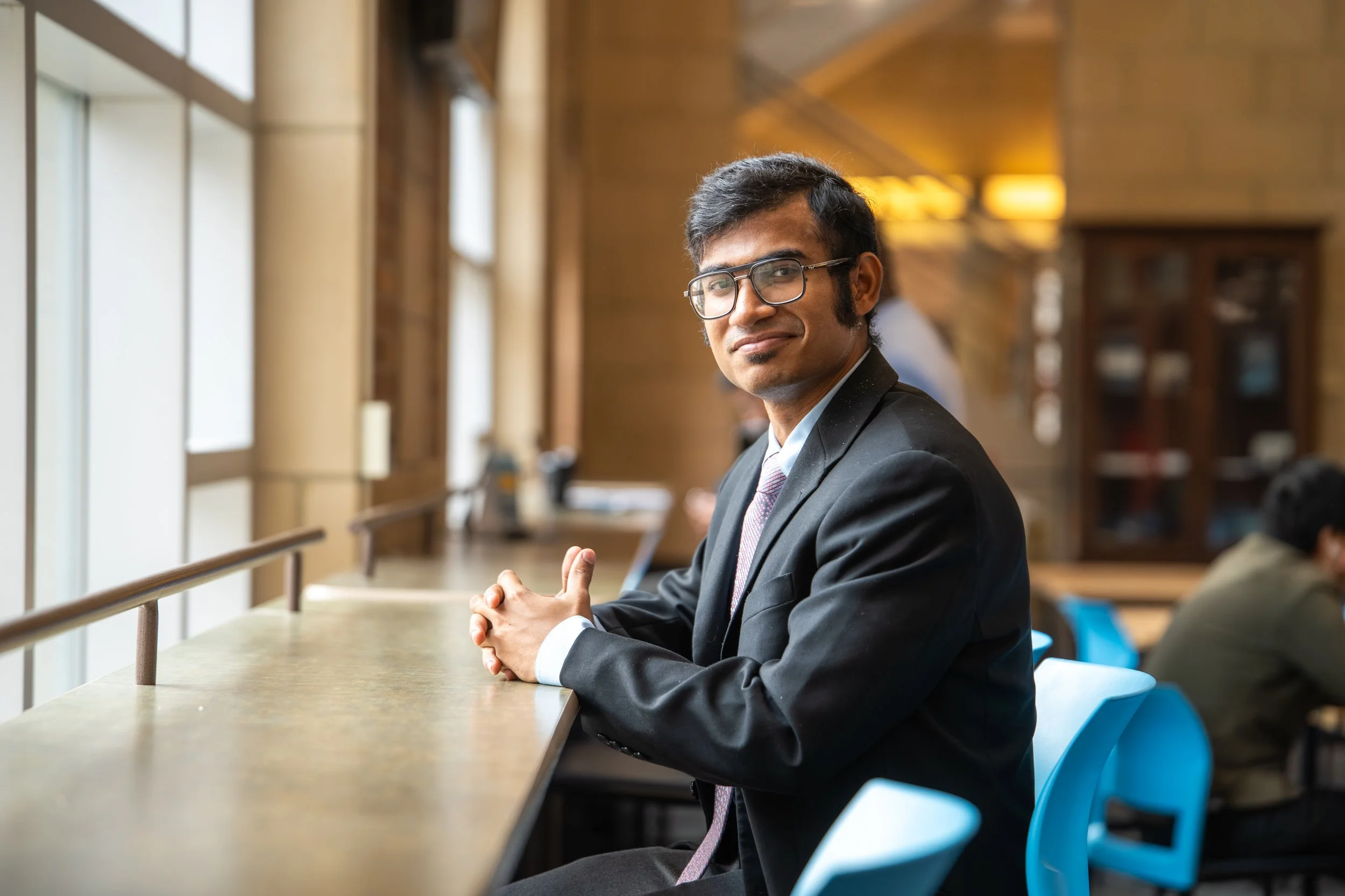 A young man in a business suit with glasses sitting at a counter in a modern, well-lit indoor space, smiling slightly and looking at the camera.