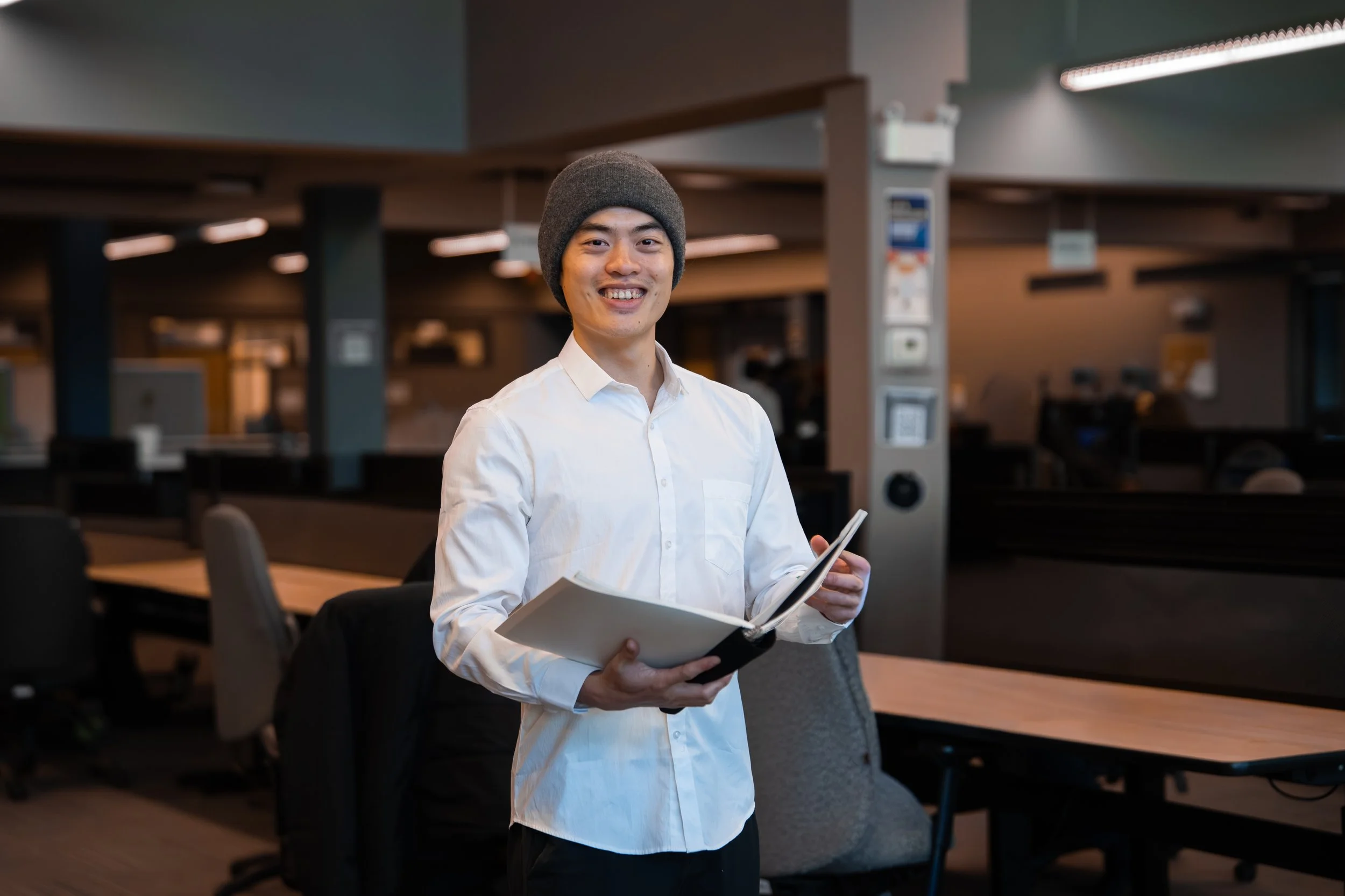 A young man with a beanie and white shirt holding a tablet and smiling in a modern office space.