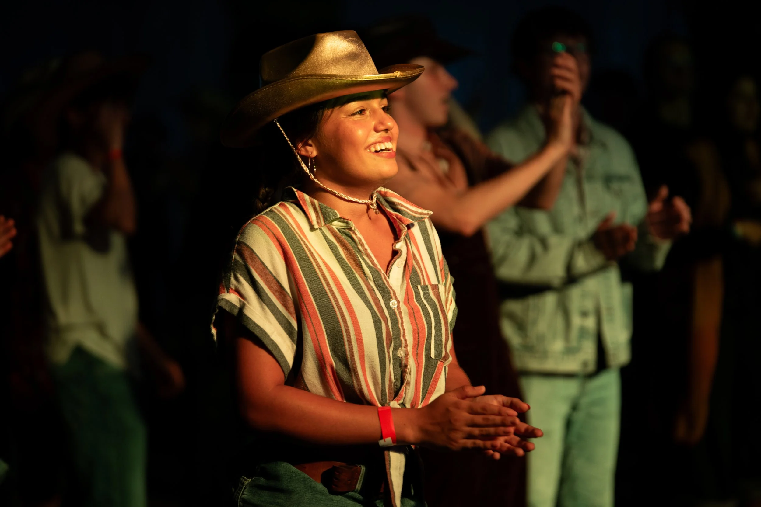 A woman with a striped shirt and a cowboy hat smiling with other people in the background, during a night event.