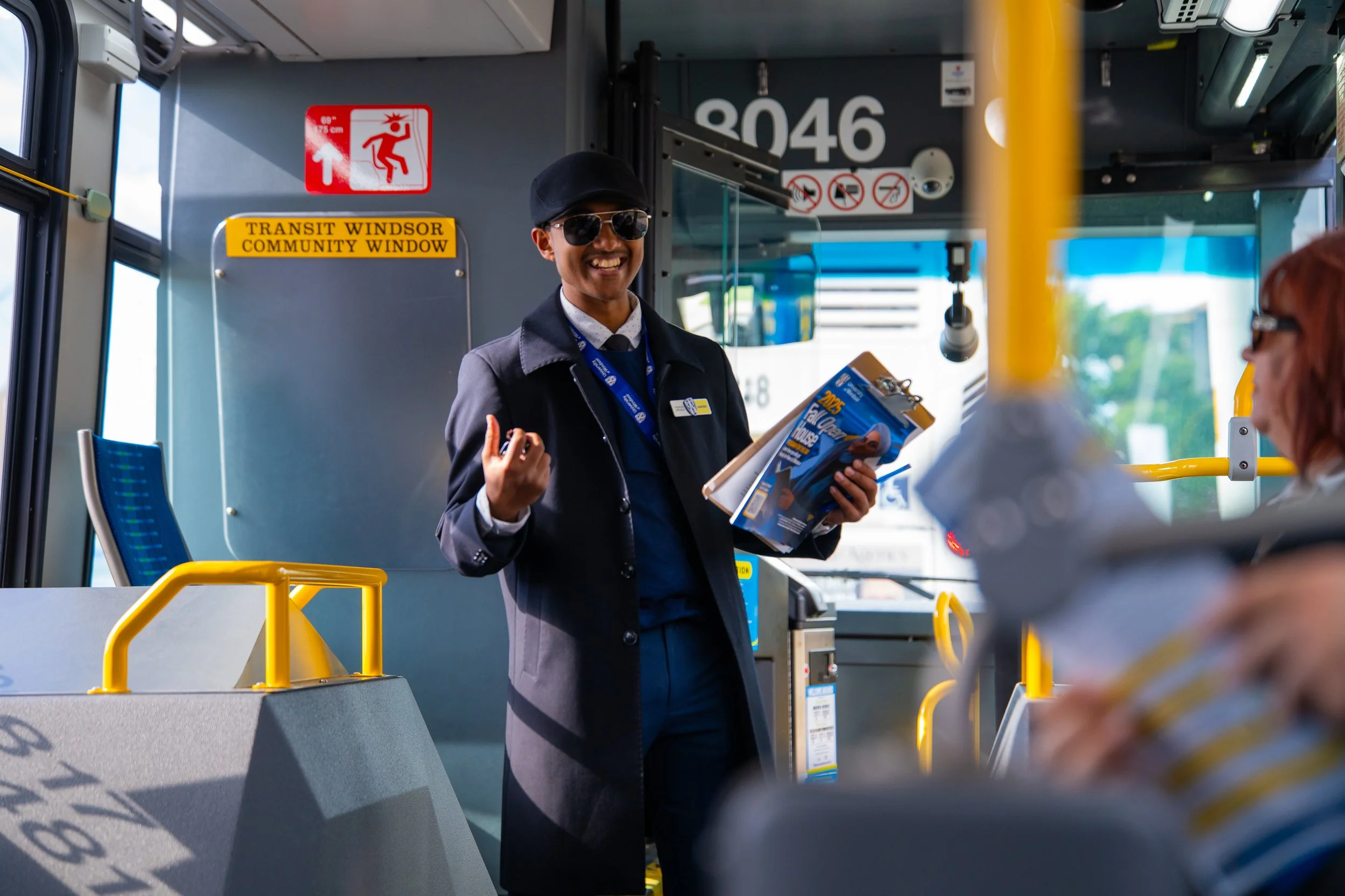 A person wearing sunglasses and a hat smiling while holding a brochure inside a bus. Visible signs include a red emergency exit sign, a yellow sign indicating the bus is part of the Transit Windsor Community Window service, and a vehicle number 3046.