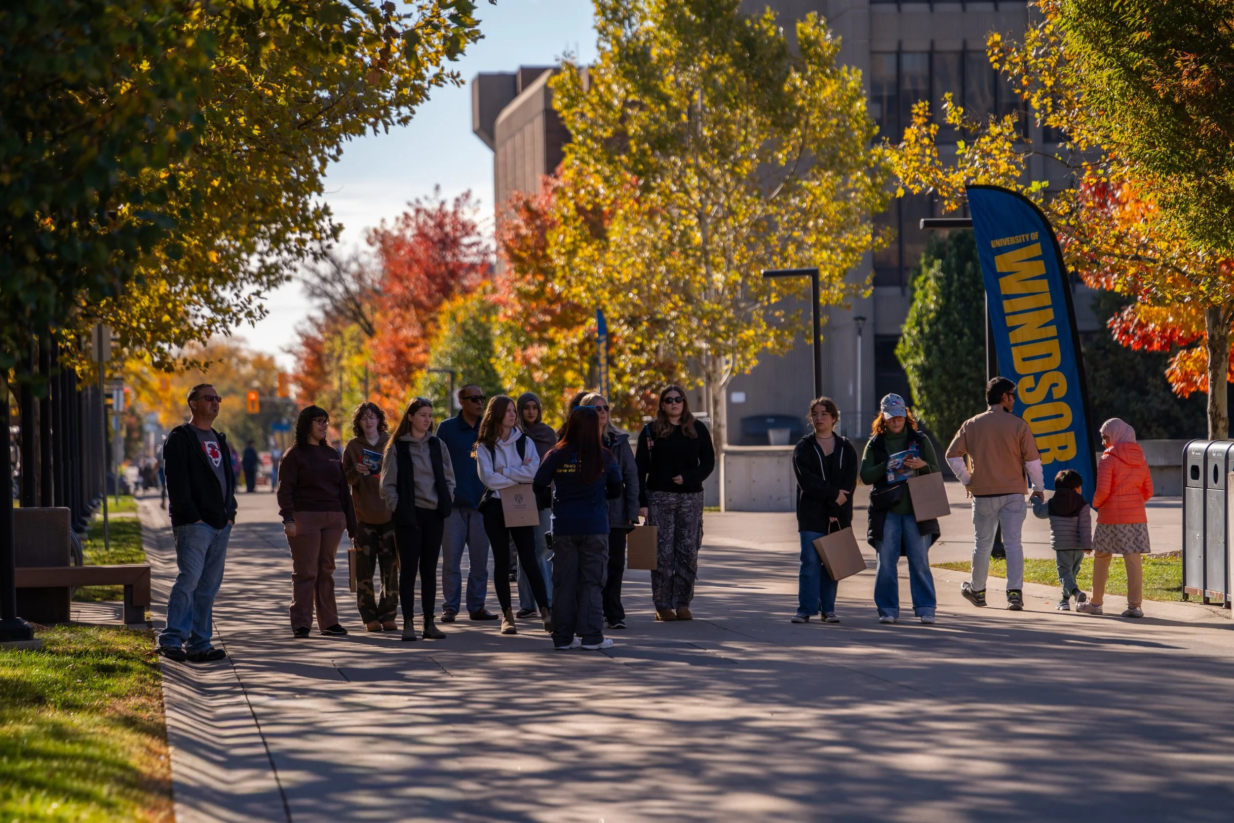 A group of people walking along a university campus sidewalk surrounded by colorful fall trees, with a blue university flag on the right.