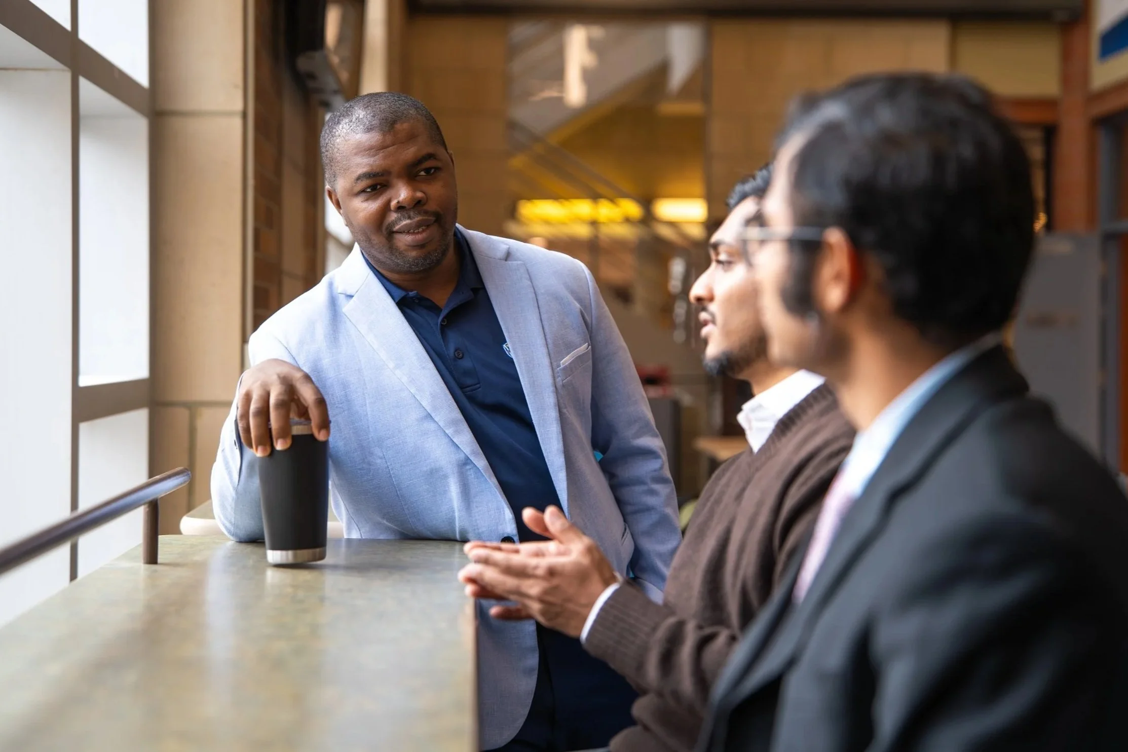 Three men engaged in conversation at a cafe or restaurant, two of them are seated and the third is standing, holding a coffee cup. The man standing is wearing a light blue blazer, the other seated man has glasses and dark hair, and the third man has dark hair and a beard, wearing a grey sweater.
