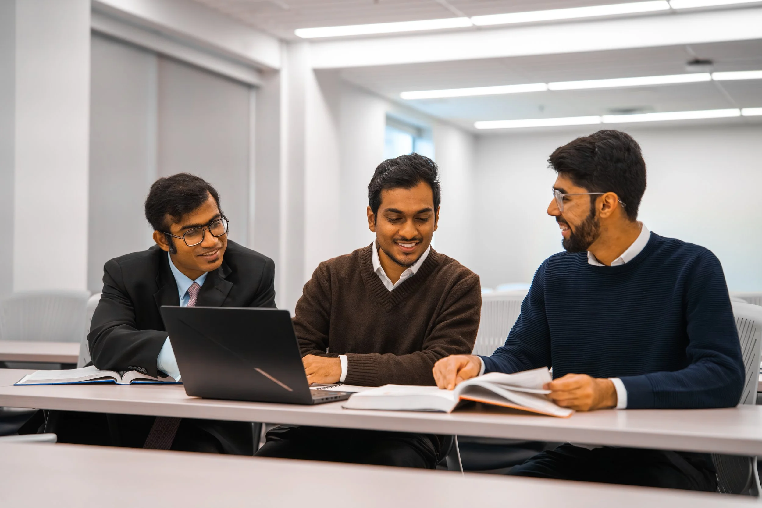 Three men in business attire sitting at a table in a modern conference room, engaged in a discussion with open books and a laptop on the table.
