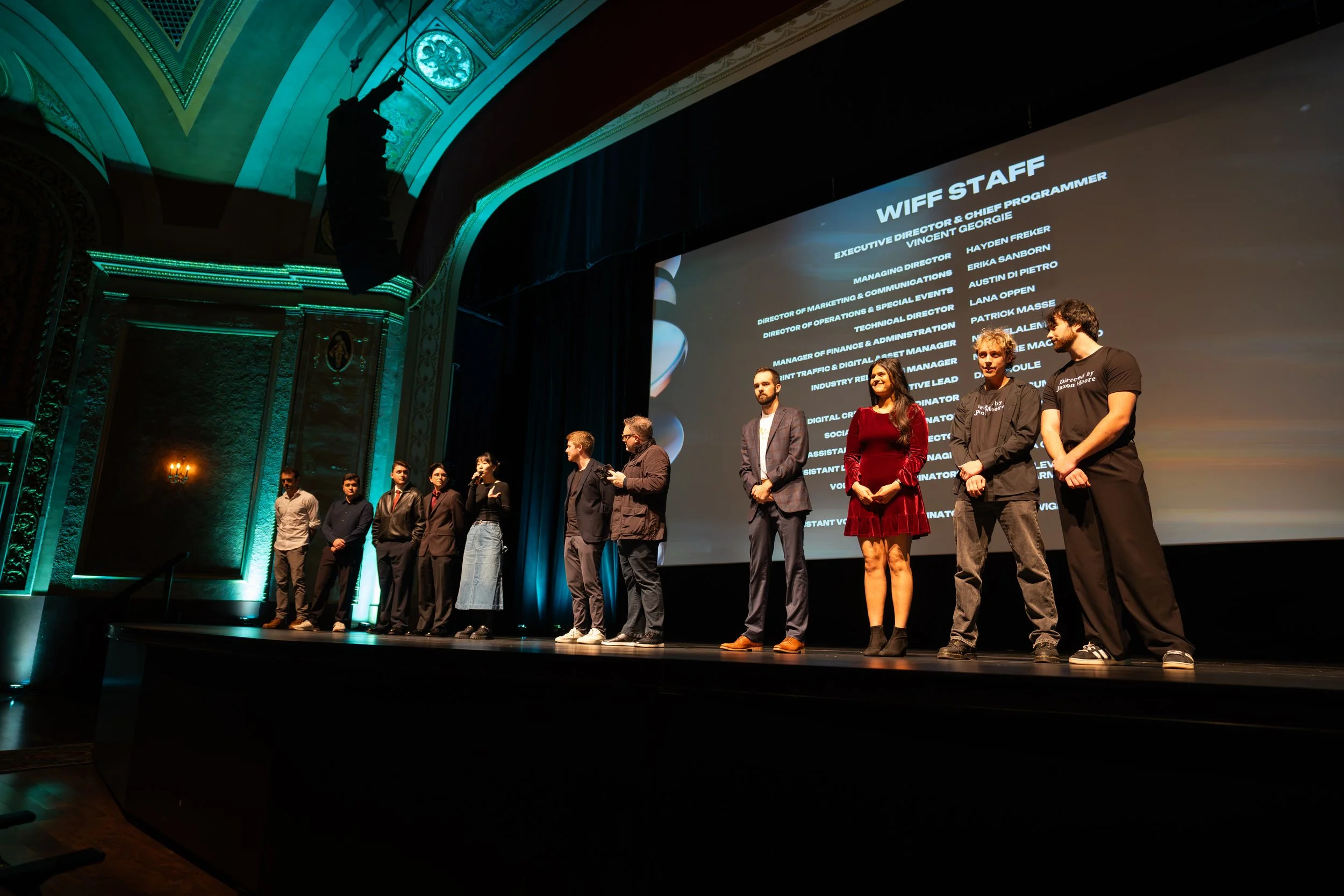 Group of people standing on stage in front of a large screen displaying the words 'WIFF STAFF' and a list of staff roles, in a theater with ornate architectural details and teal lighting.