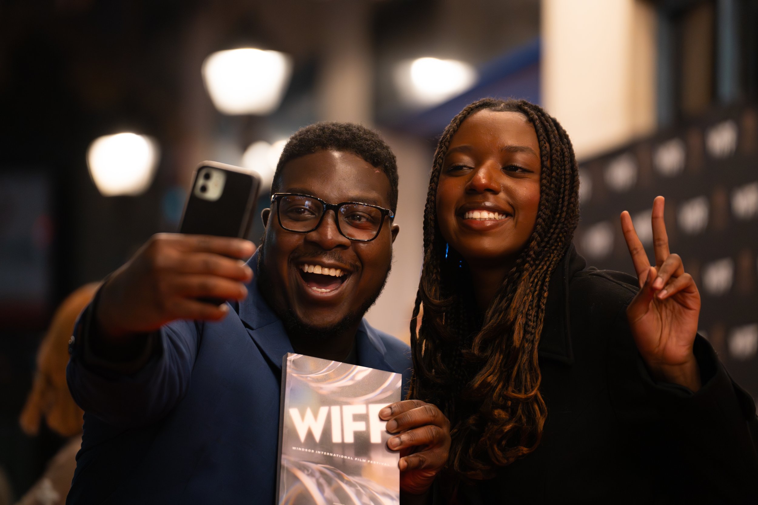 Two smiling people taking a selfie at a festival, with one person holding a brochure or program.